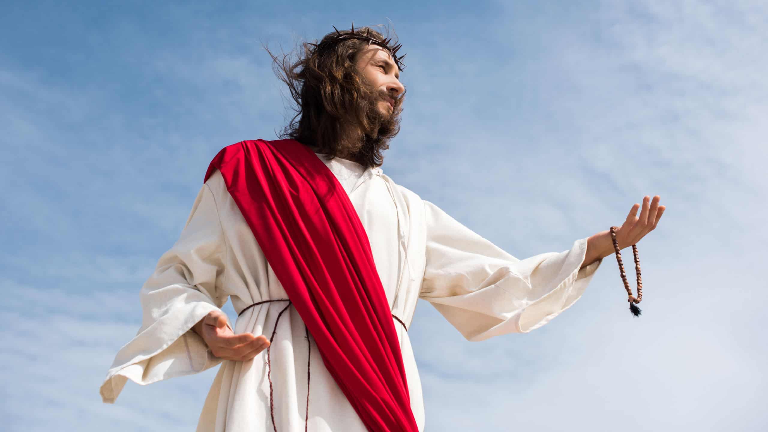 low angle view of Jesus in robe holding rosary and reaching hand against blue sky