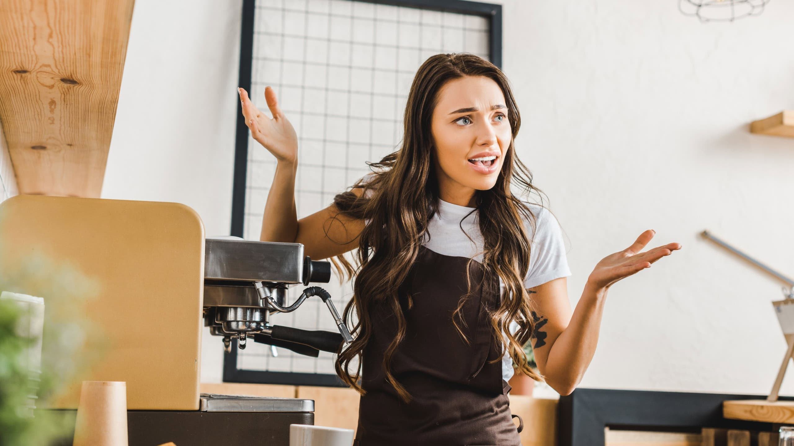 angry brunette barista in brown apron standing near coffee machine in coffee house