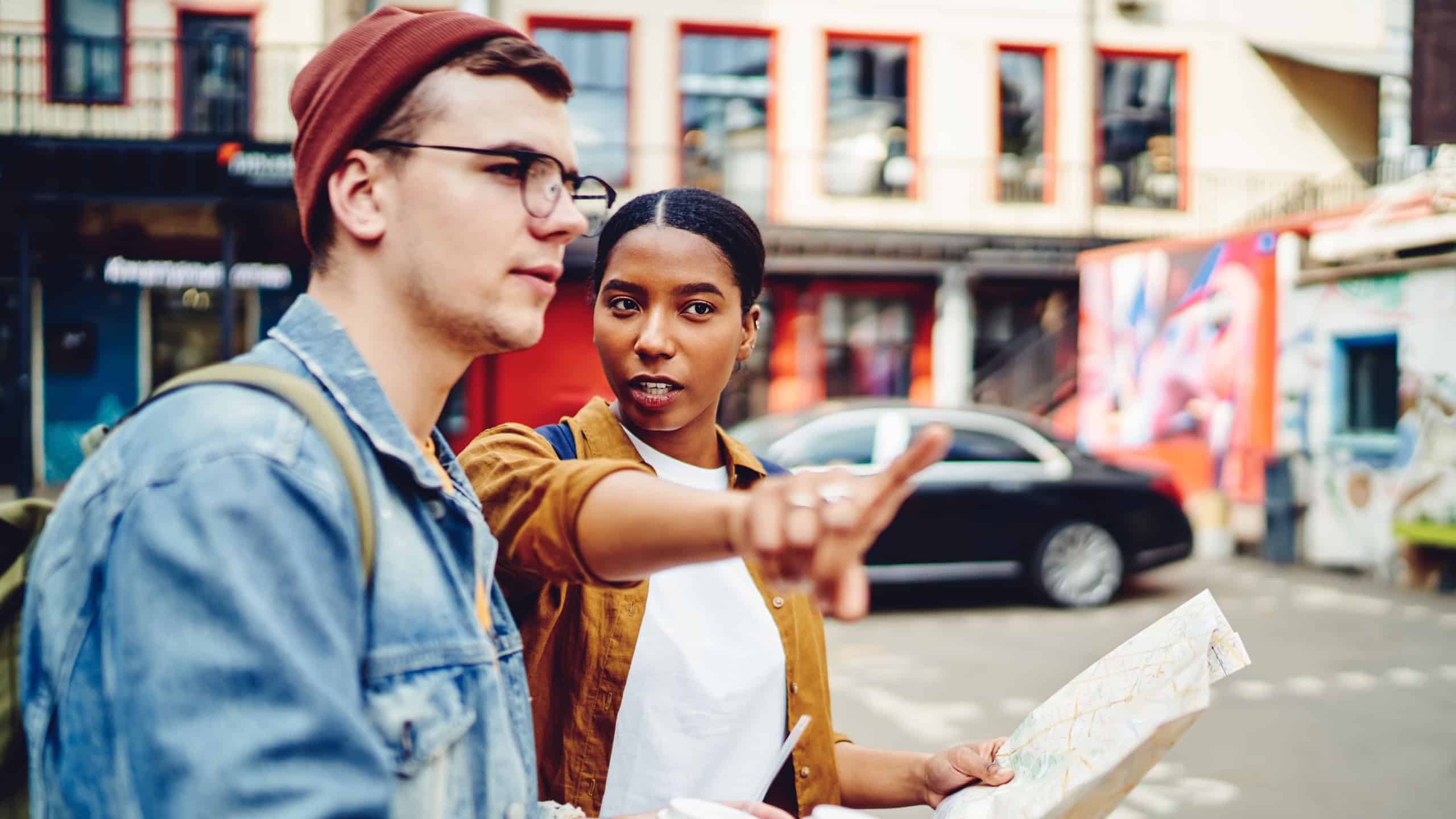 African American young woman inhabitant of city pointing hand on right direction to help caucasian tourist with map to find destination. Male traveler walking on street and search showplace in town