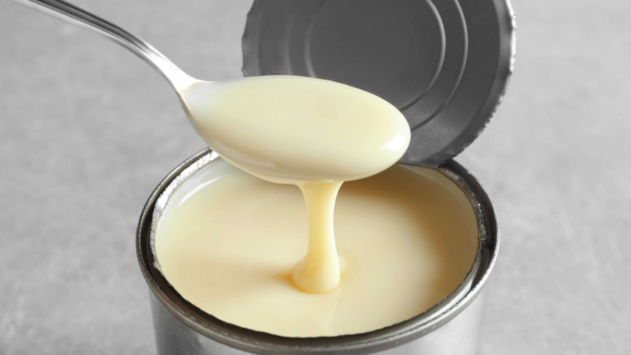Condensed milk pouring from spoon into tin can on grey background, closeup. Dairy product