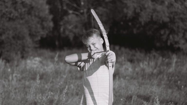 Cute caucasian young child playing wooden toy bow and arrow outside. Setting goals concept. Horizontal black and white photography.
