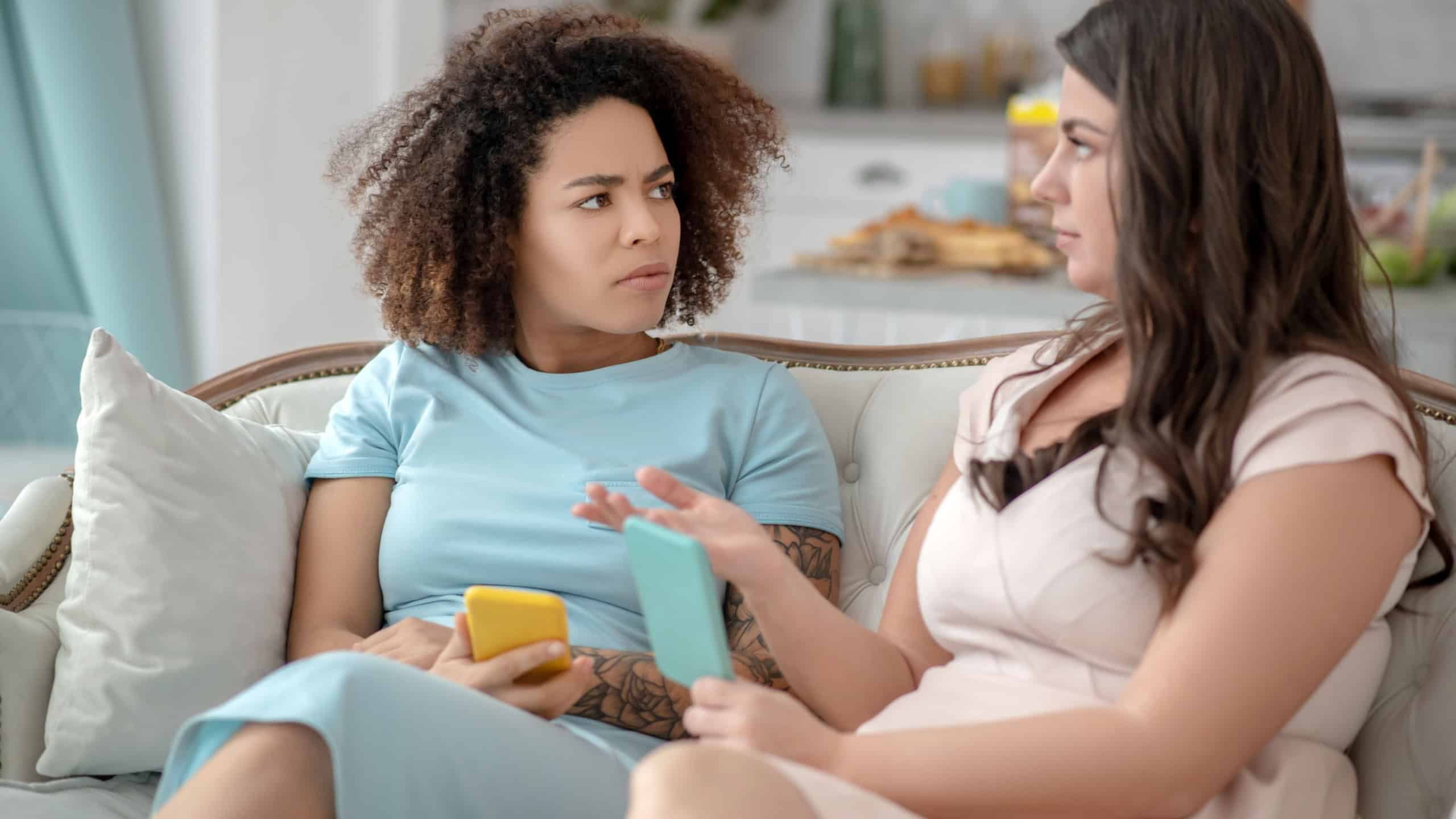 Easy conversation. Serious woman in a light blue dress with a yellow smartphone, calm gesticulating in a beige dress with a mint smartphone, chatting on the couch.