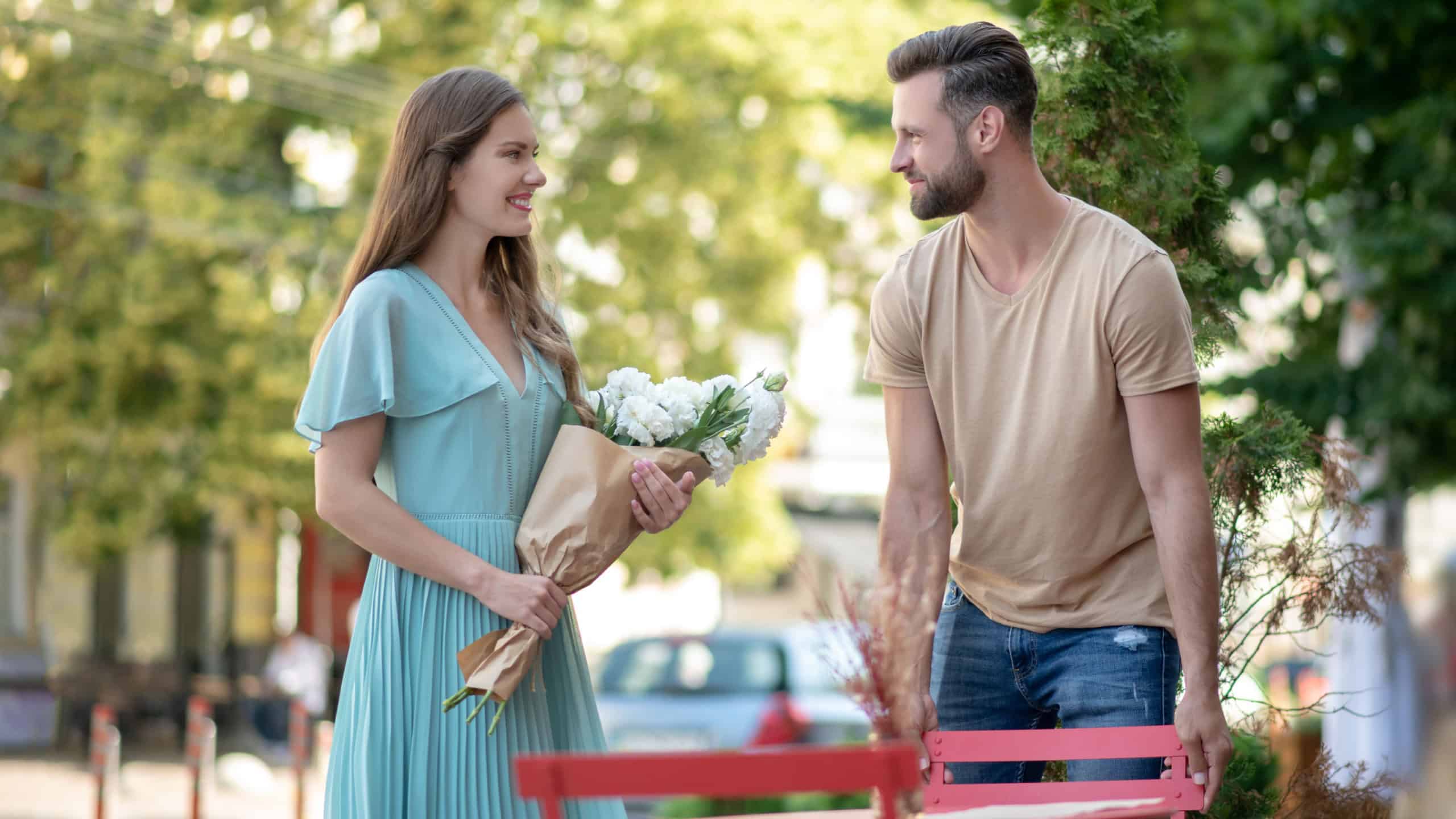 Lovely date. Bearded male pulling red chair for cute young female with bouquet of flowers