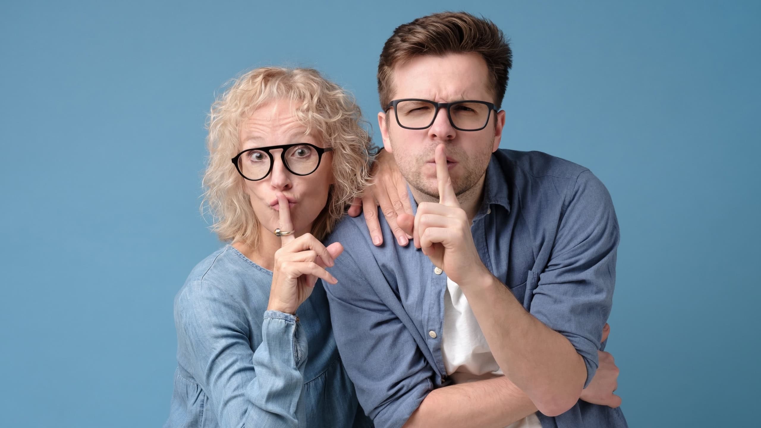 Mother and son holding fingers on lips mouth to keep it quiet, hush secret showing shh shush gesture for conspiracy. Studio shot on blue wall.