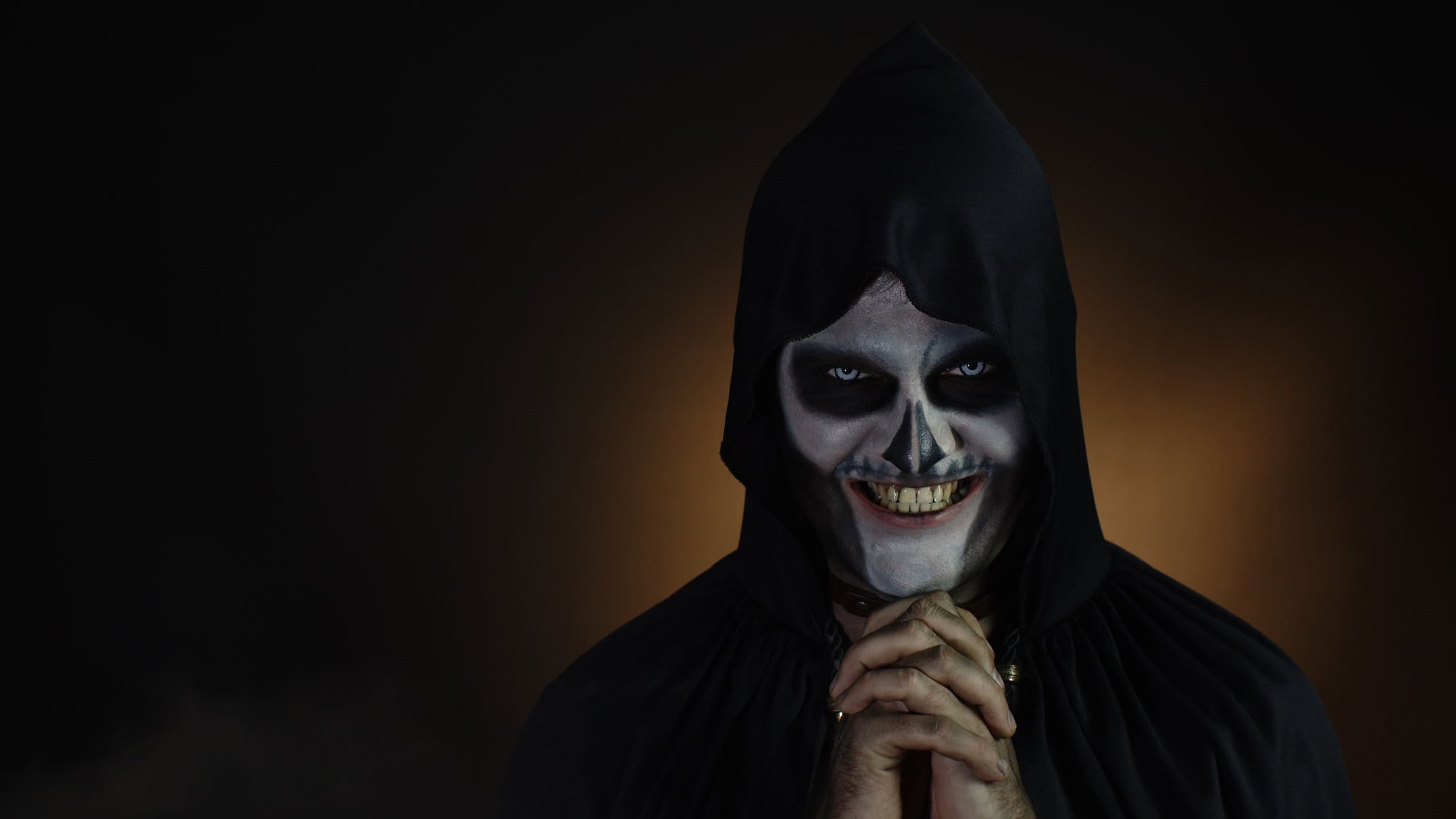 Creepy, crazy man with skeleton makeup in hood. Guy executioner making angry faces, shaking head, trying to scare. Voodoo rituals. Baron Saturday. Halloween party. Black background