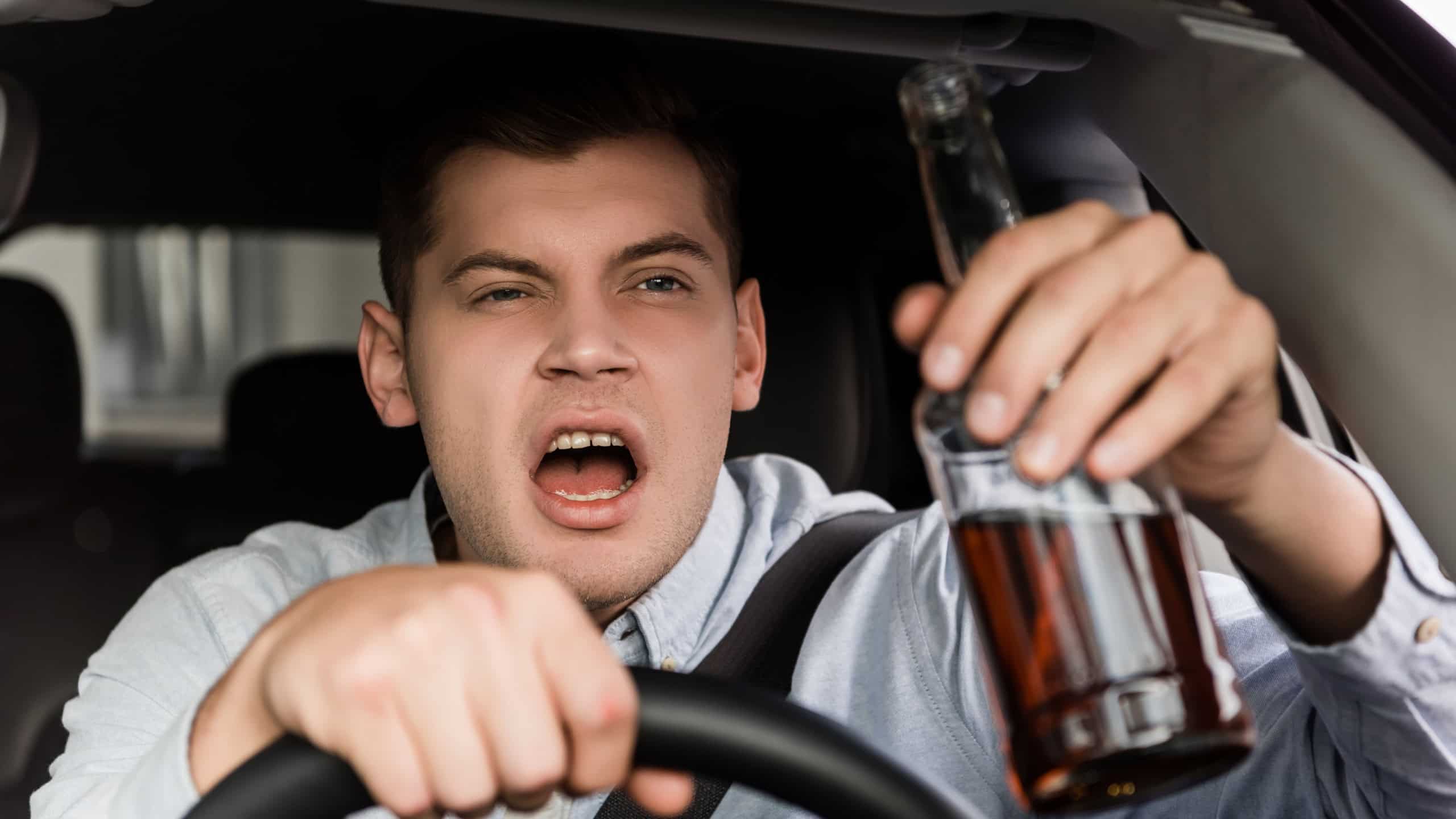 Drunk, aggressive man shouting while driving car and holding bottle of alcohol on blurred foreground