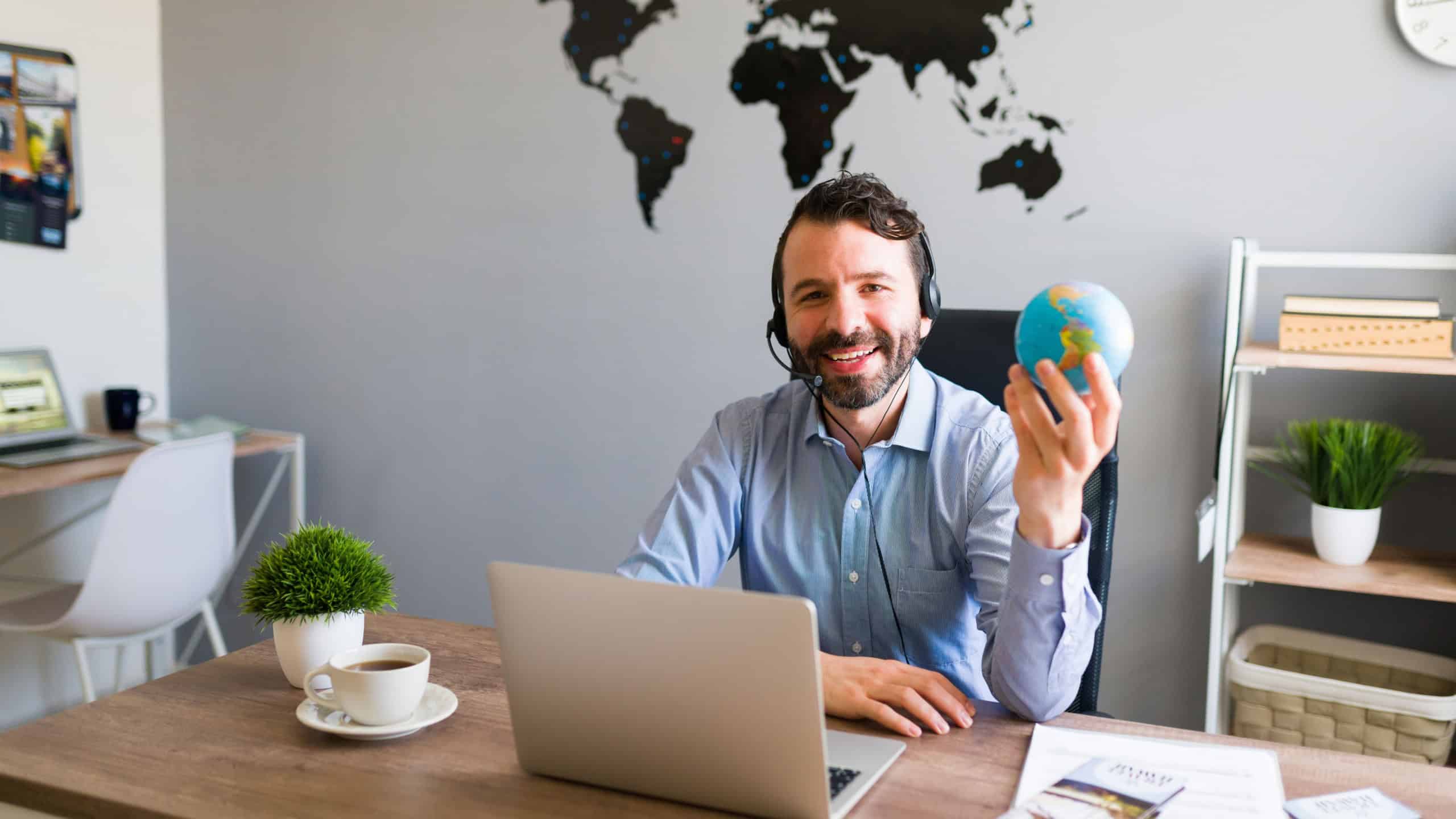 I can help you with your next vacation trip. Portrait of a good-looking man and sales agent holding a globe at his travel office with a world map