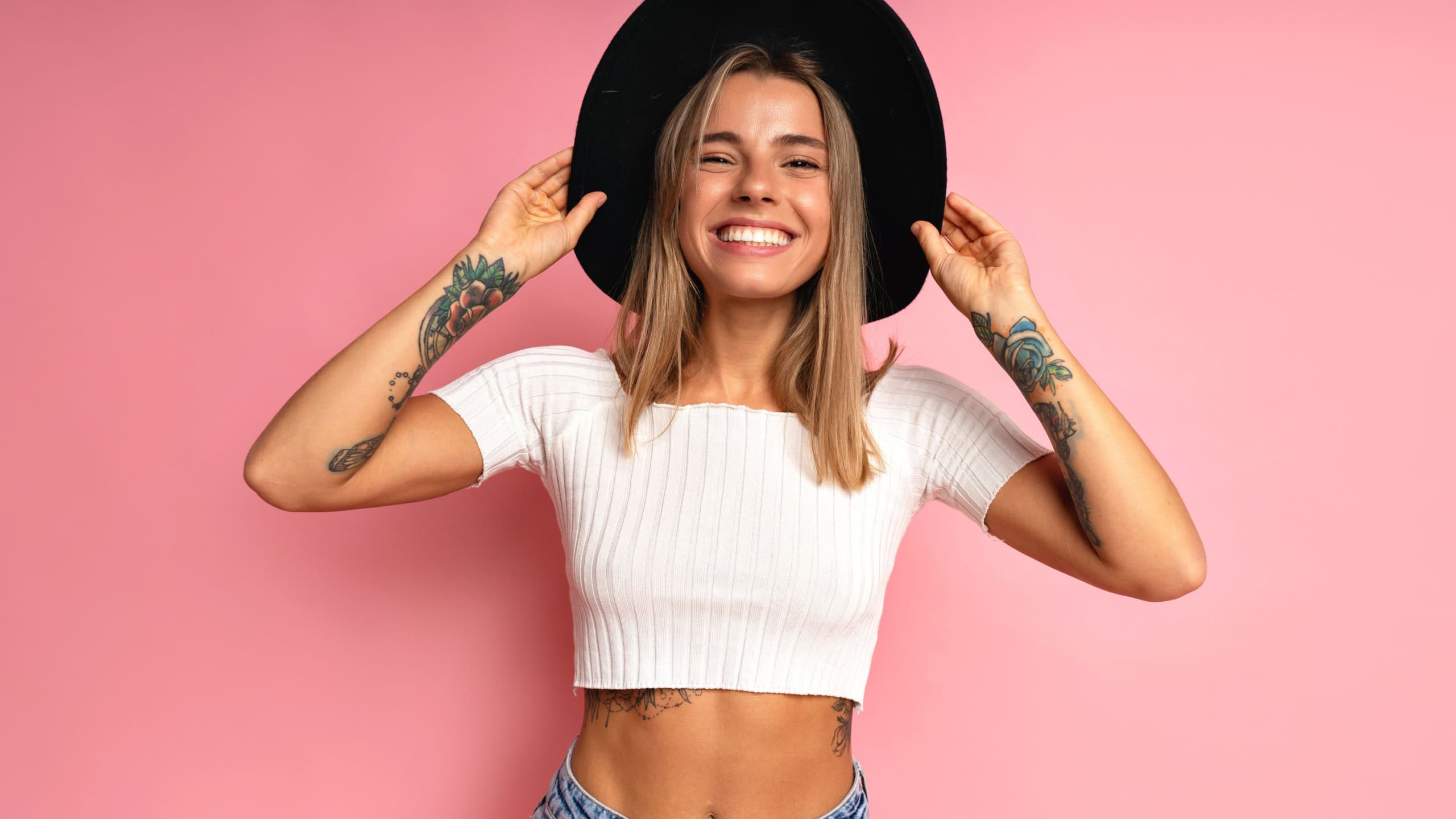 Cheerful stylish woman posing at studio pink background, wearing crop top blue jeans, black fedora. Blonde hairs and tattos.