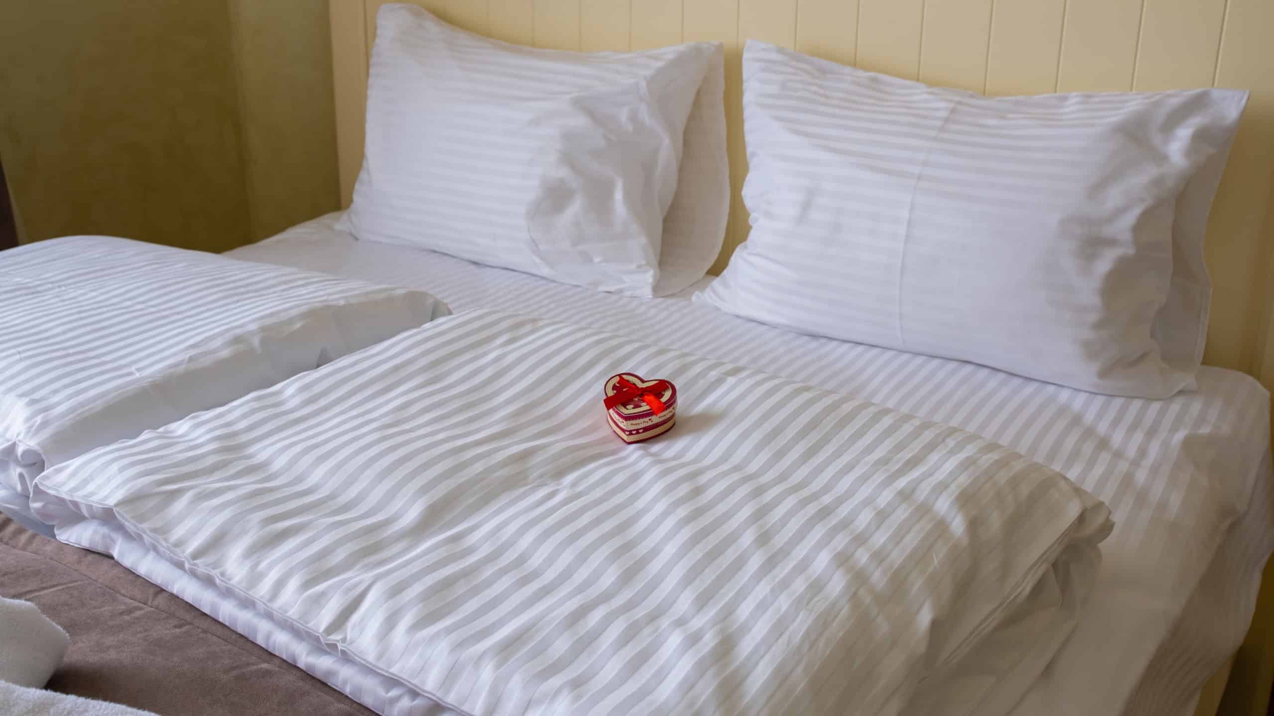 A small gift box in the shape of a heart on a white bed in a hotel room.