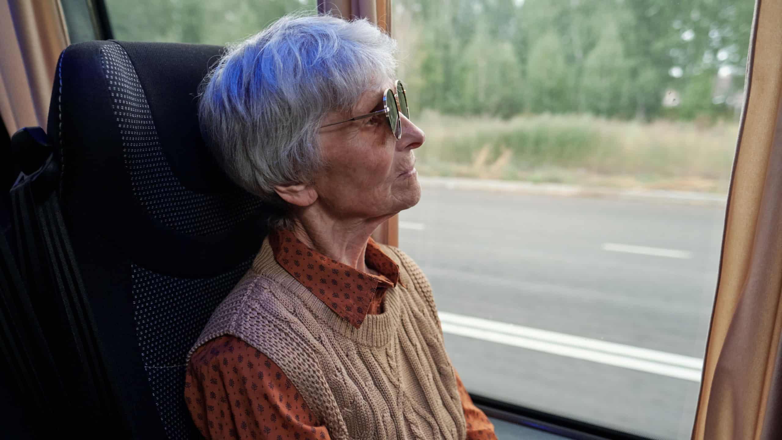 Serious senior Caucasian woman in sunglasses looking through window while riding bus