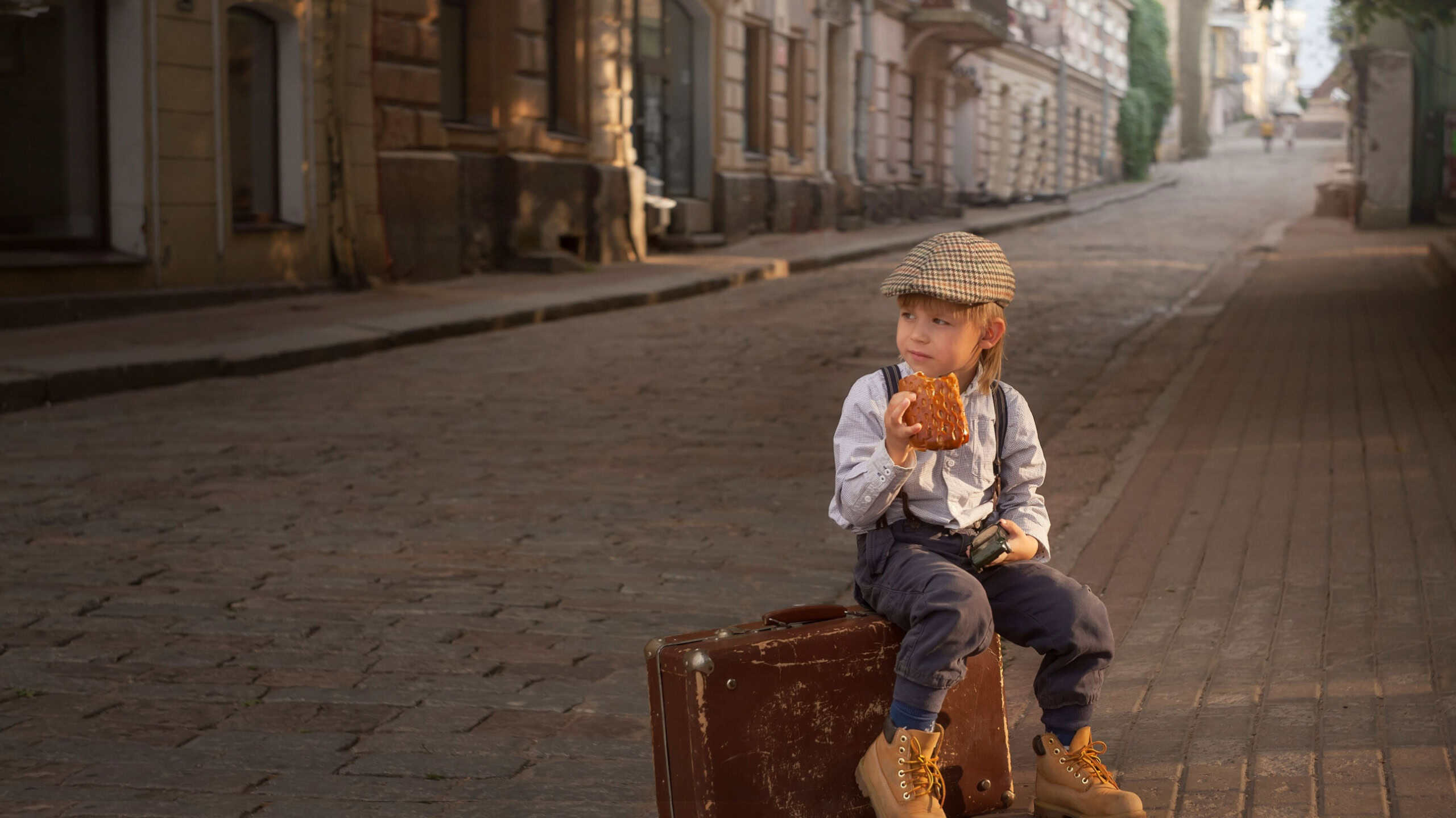 A boy in vintage clothes is sitting on a suitcase by the road and eating a bun. He is wearing a tweed cap, rough boots, a shirt and suspenders.