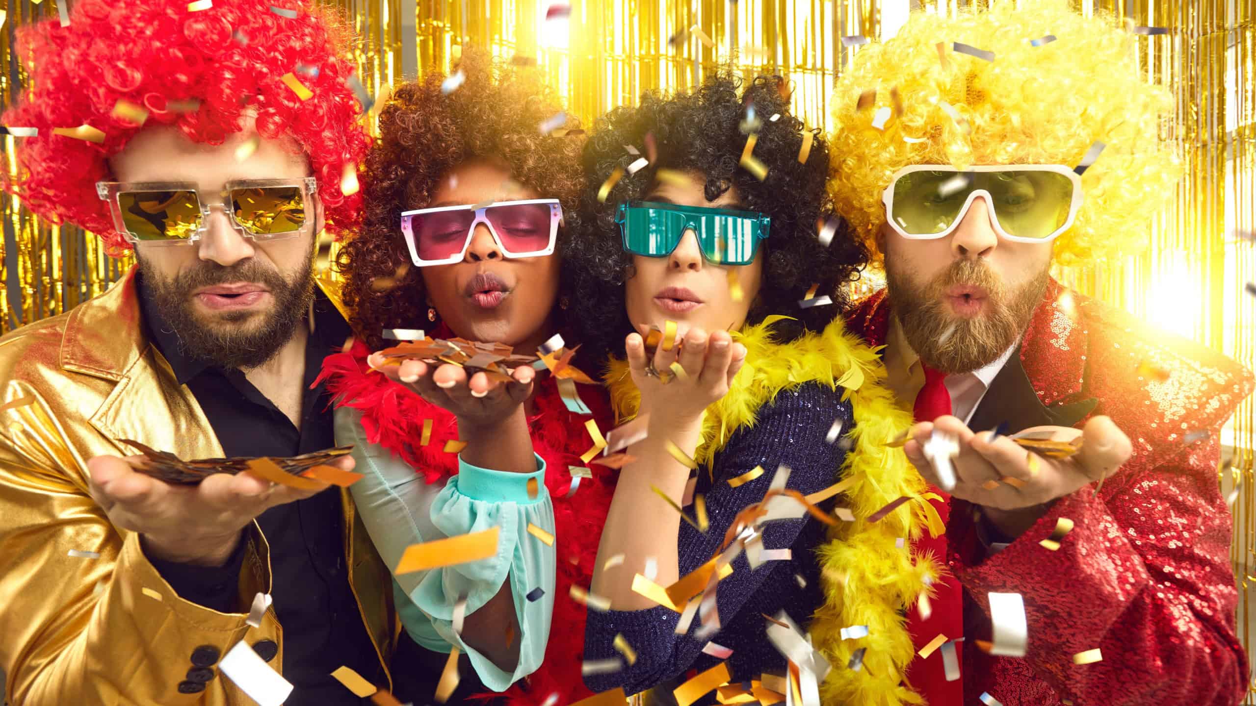 Happy merry diverse people dressed in boas, glasses and curly Afro wigs celebrating New Year at disco night club party throw a kiss blowing shiny golden confetti sharing groovy festive mood