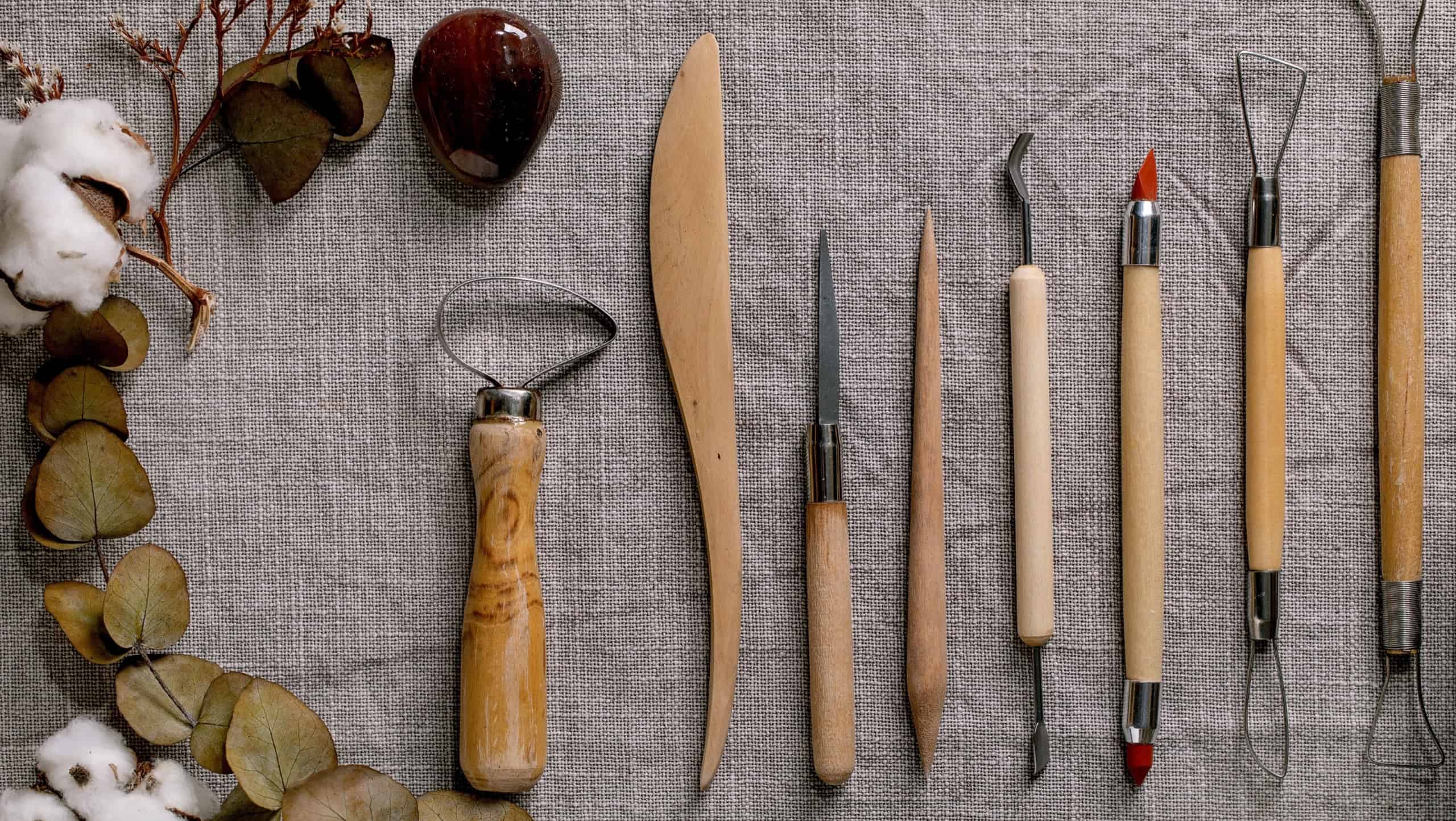 Set for pottery hobby. Kit of pottery ceramics tools in row on linen fabric cloth with cotton flowers and eucalyptus. Top view. DIY potter hobby