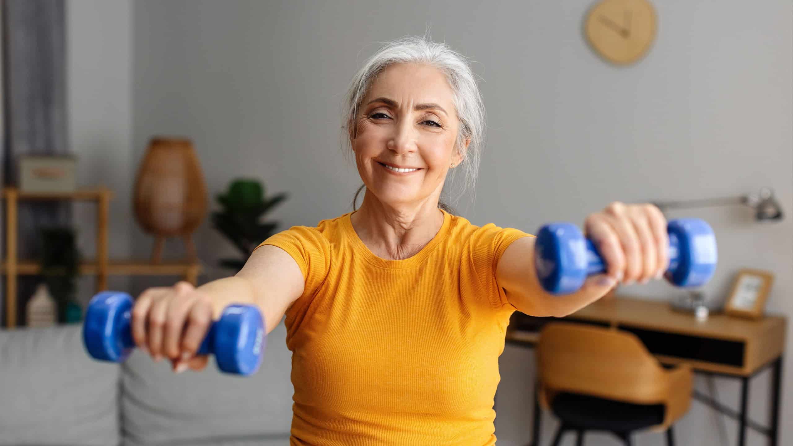 Health care, sport and active lifestyle. Happy senior woman doing exercises with dumbbells in living room interior, looking at camera and smiling