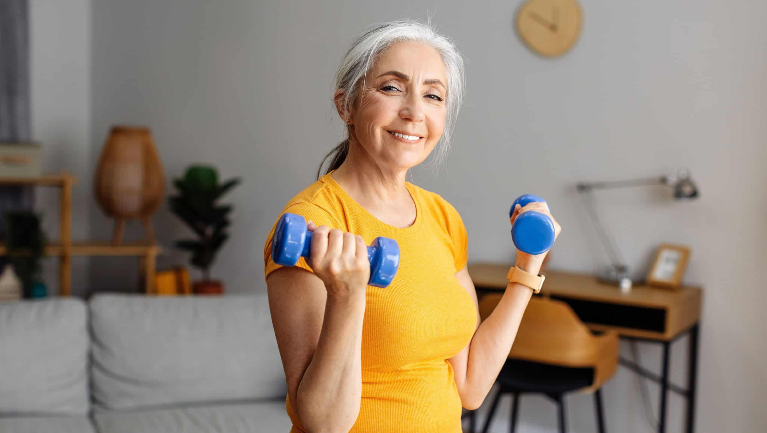 Happy senior woman doing dumbbell workout at home, working on arms strength, looking at camera and smiling. Fit aged lady lifting fitness dumbells up over living room interior