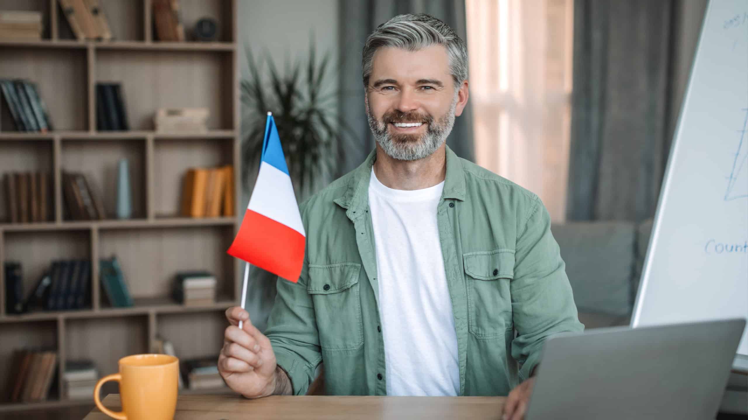 Glad senior caucasian man teacher with beard hold flag of France near blackboard in room interior.