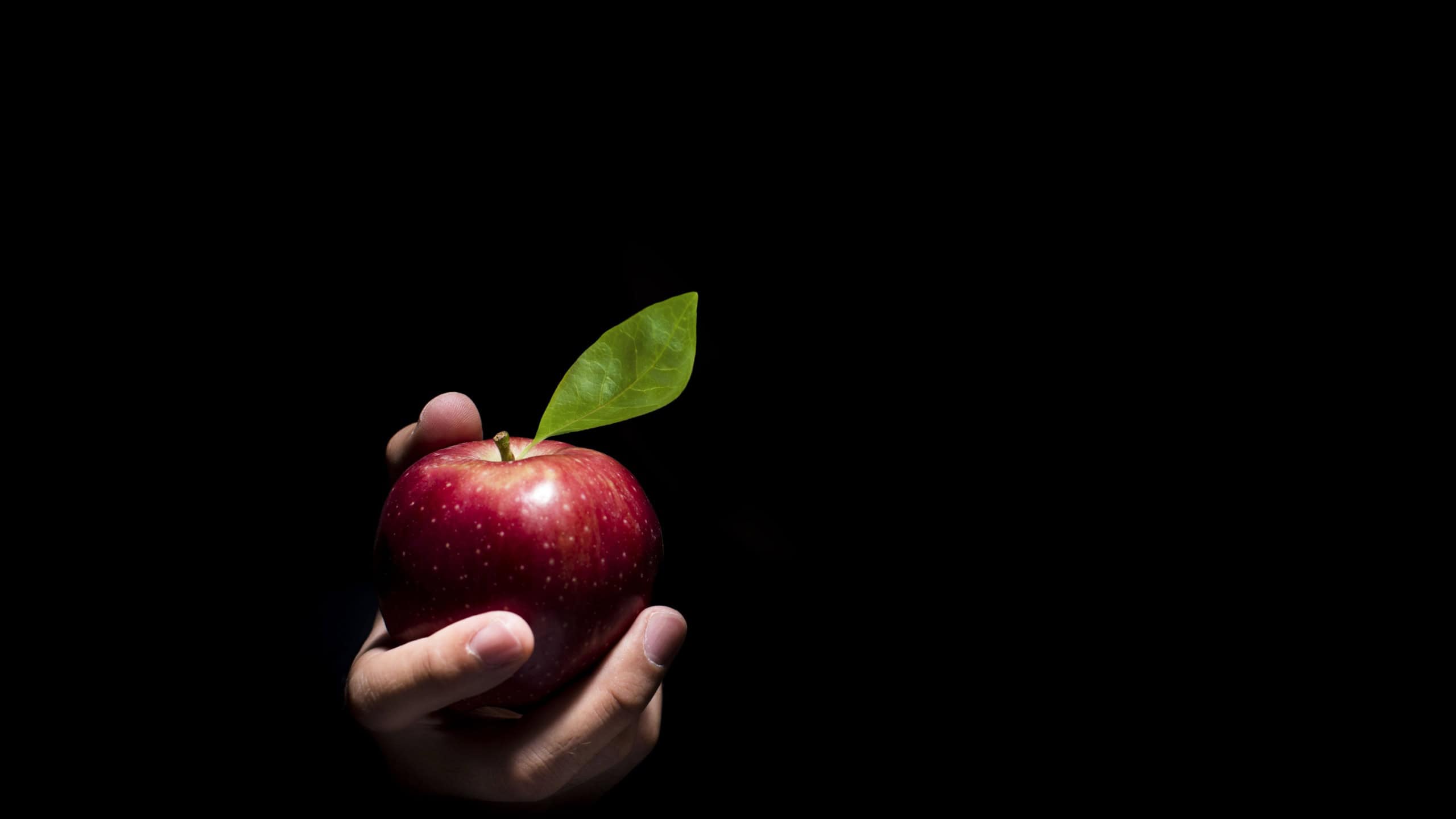 Hand offering a red apple on a black background.