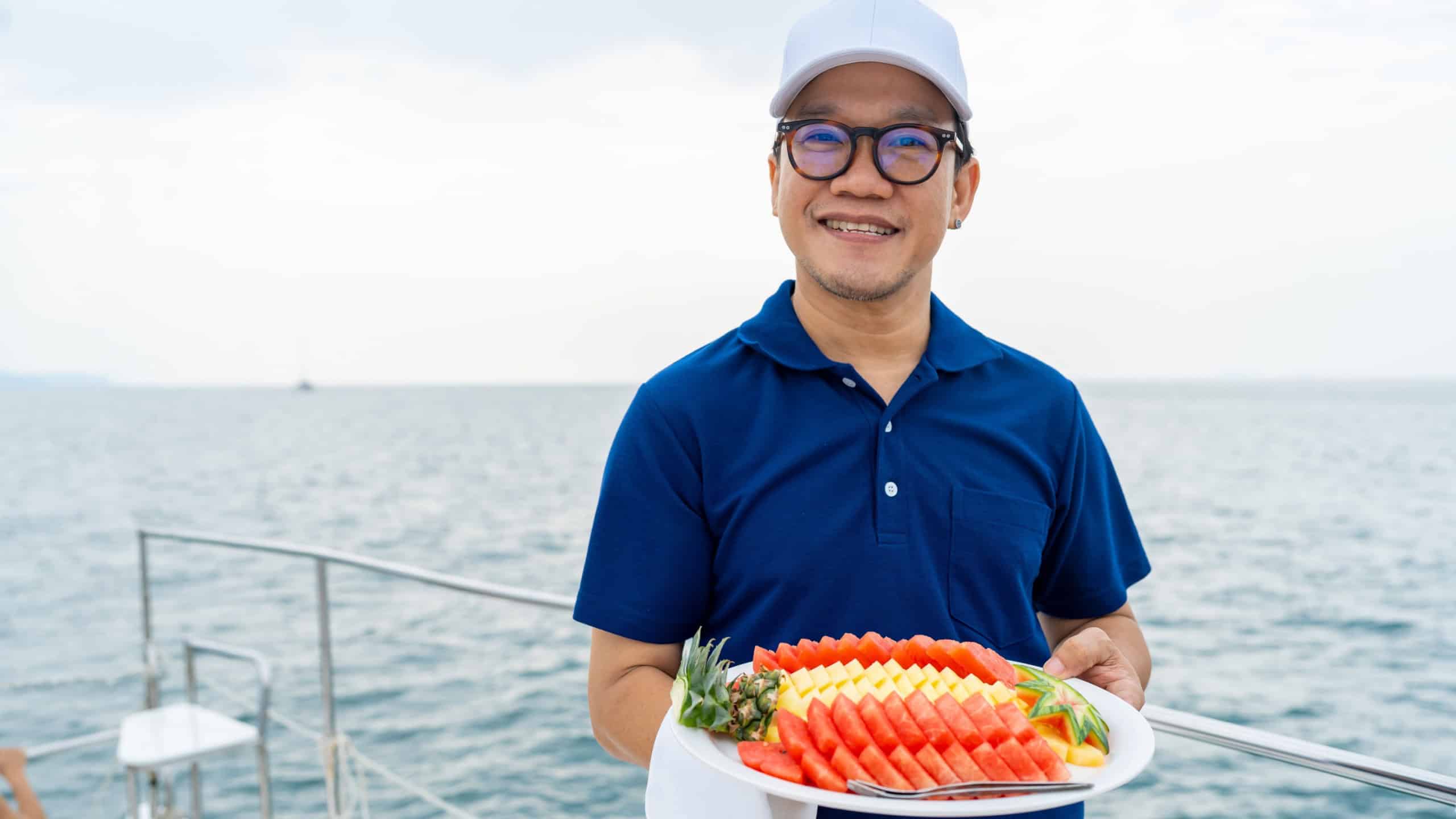 ortrait of Happy Asian man waiter holding fresh fruit on serving dish for serving to passenger tourist travel on cruise