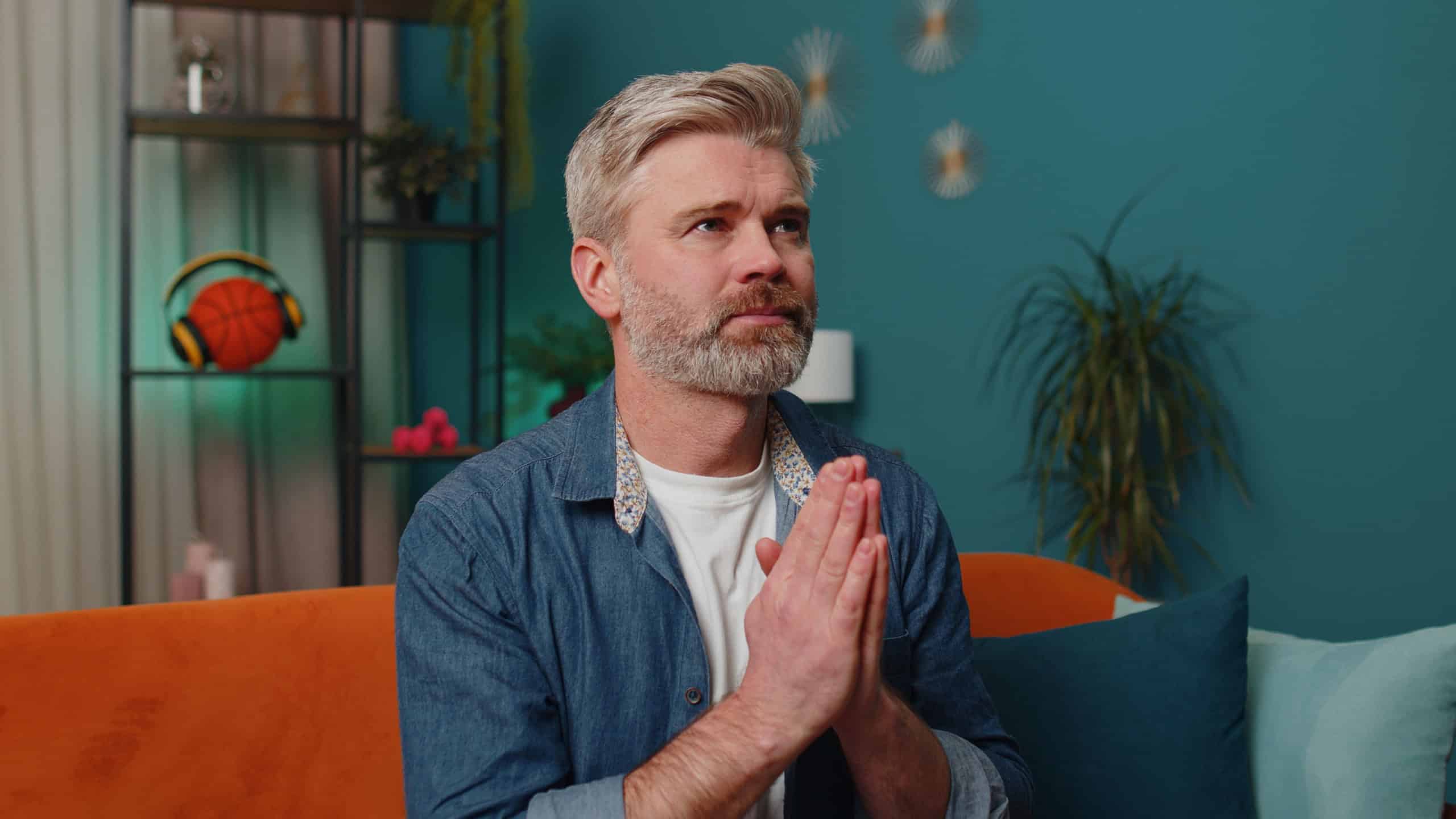 Please, God help. Middle-aged man praying sincerely with folded arms, looking upward and making wish, asking with hopeful imploring expression, begging apology. Mature senior gray-haired guy at home