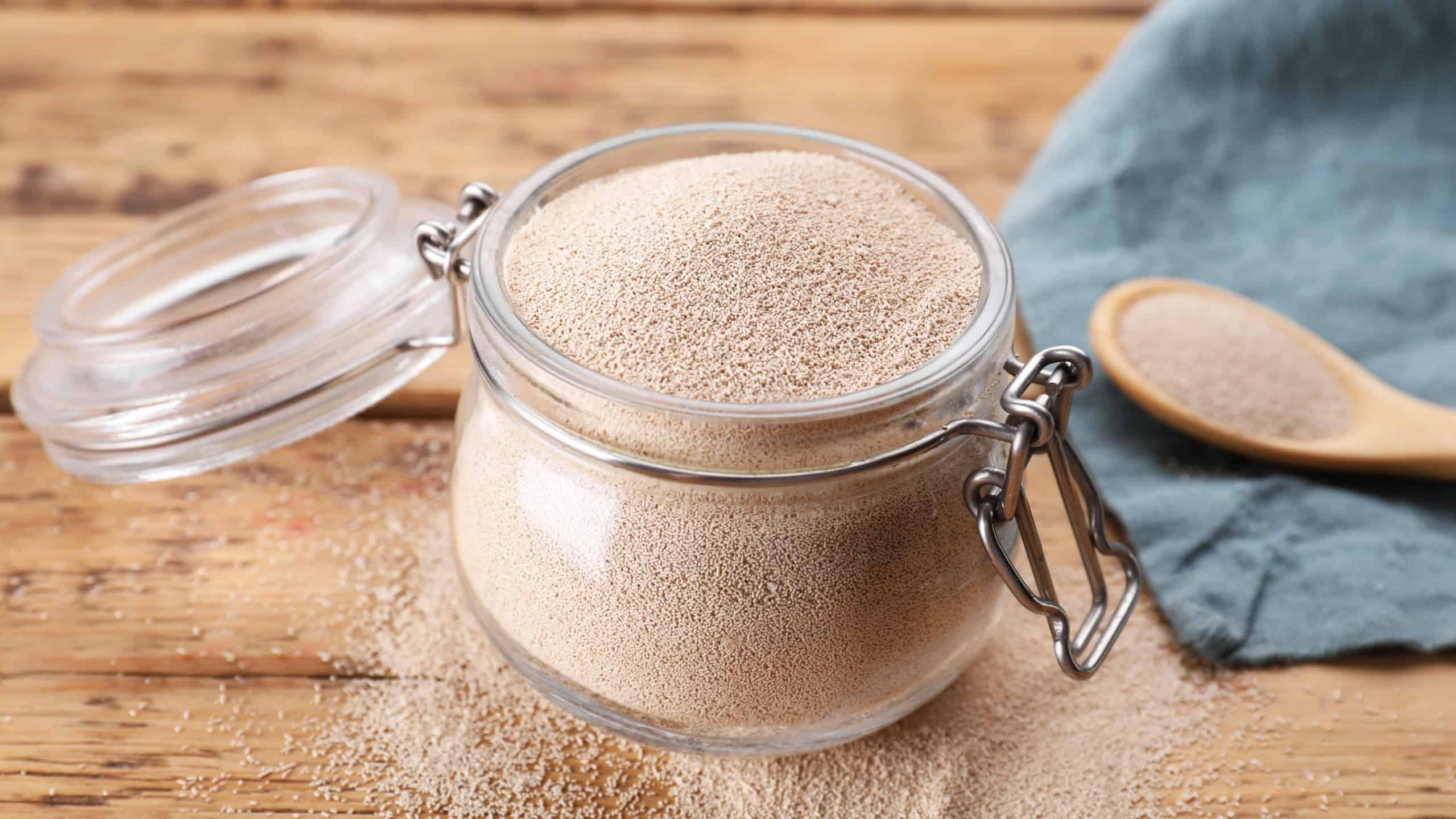 Glass jar and spoon with active dry yeast on wooden table, closeup