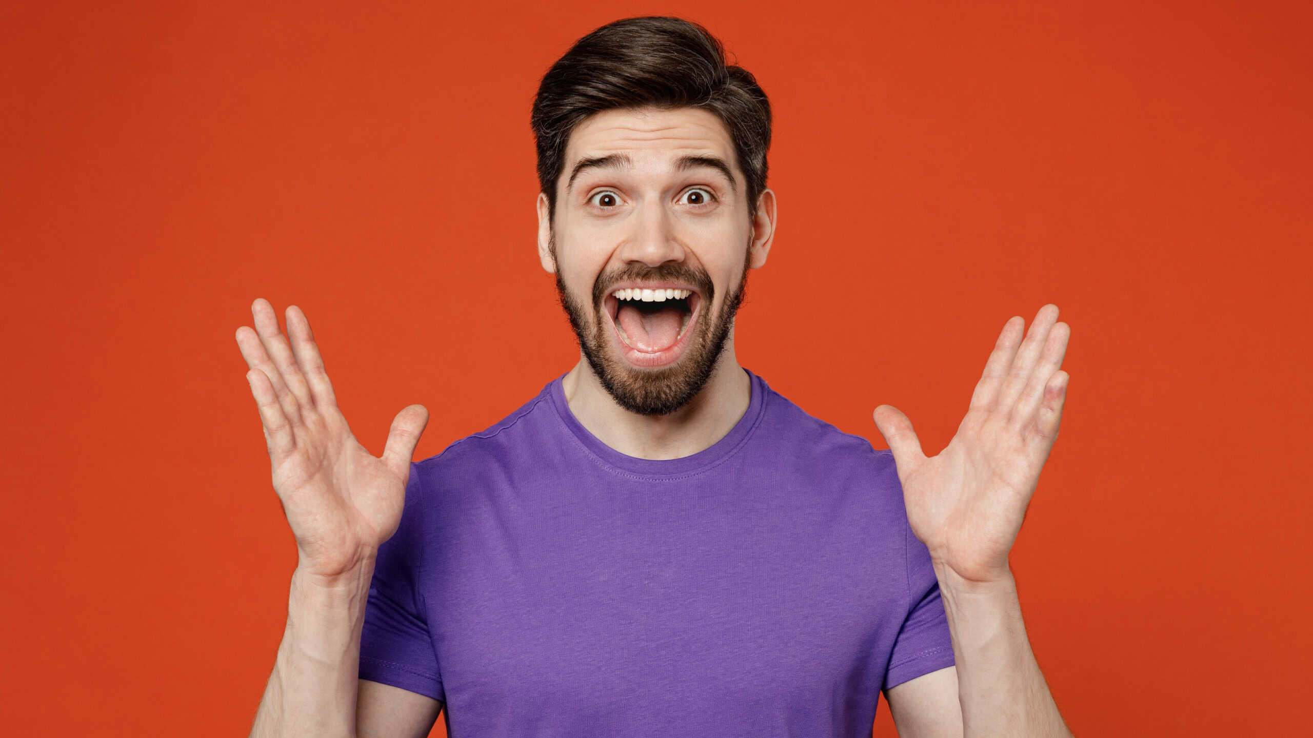 Young surprised shocked happy brunet caucasian man 30s wear casual basic purple t-shirt look camera spread hands say wow isolated on plain orange background studio portrait. People lifestyle concept