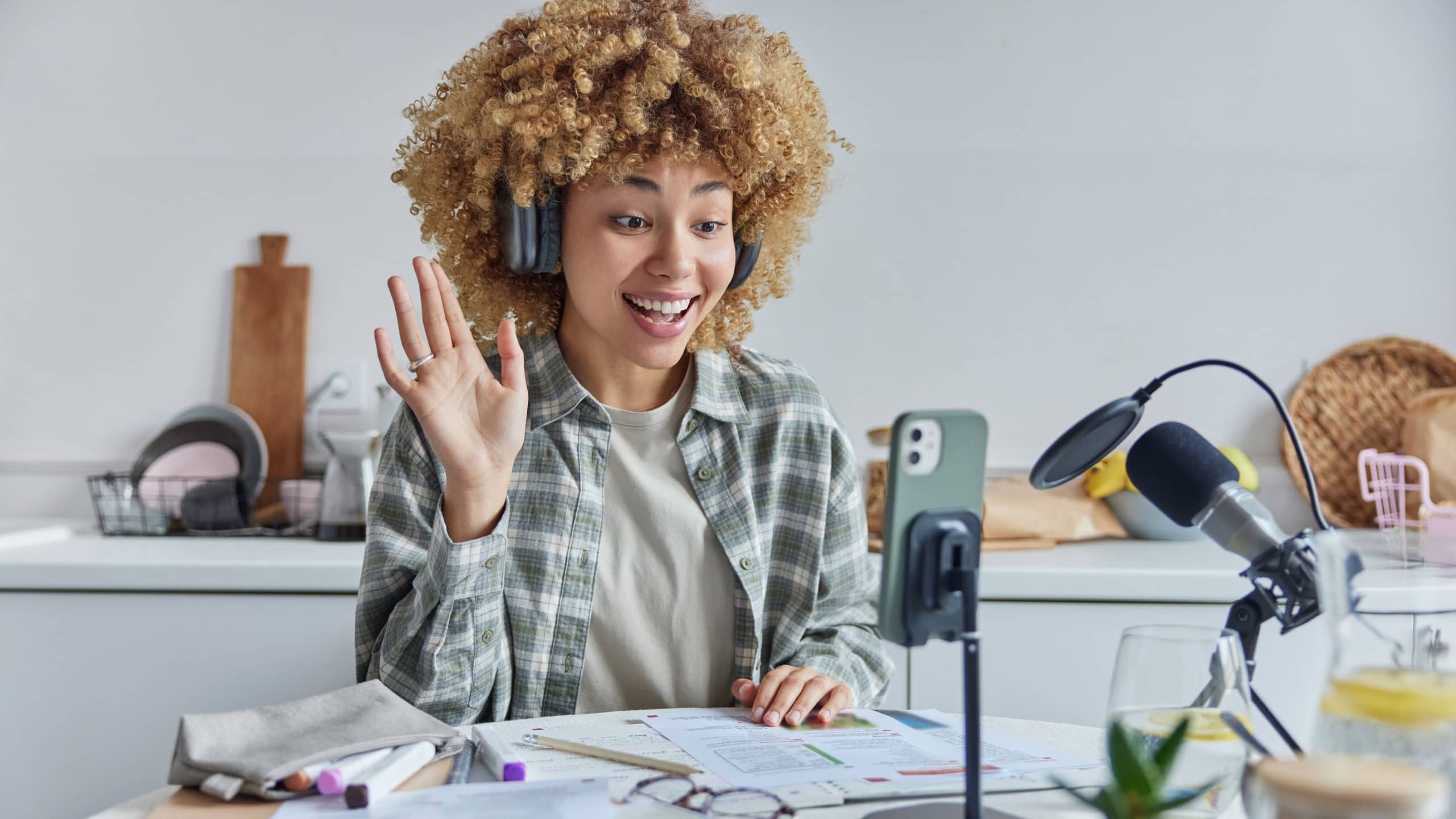 Online education home schooling concept. Positive curly haired female student waves hello to her tutor has distance lesson uses modern technologies high speed internet poses against kitchen interior