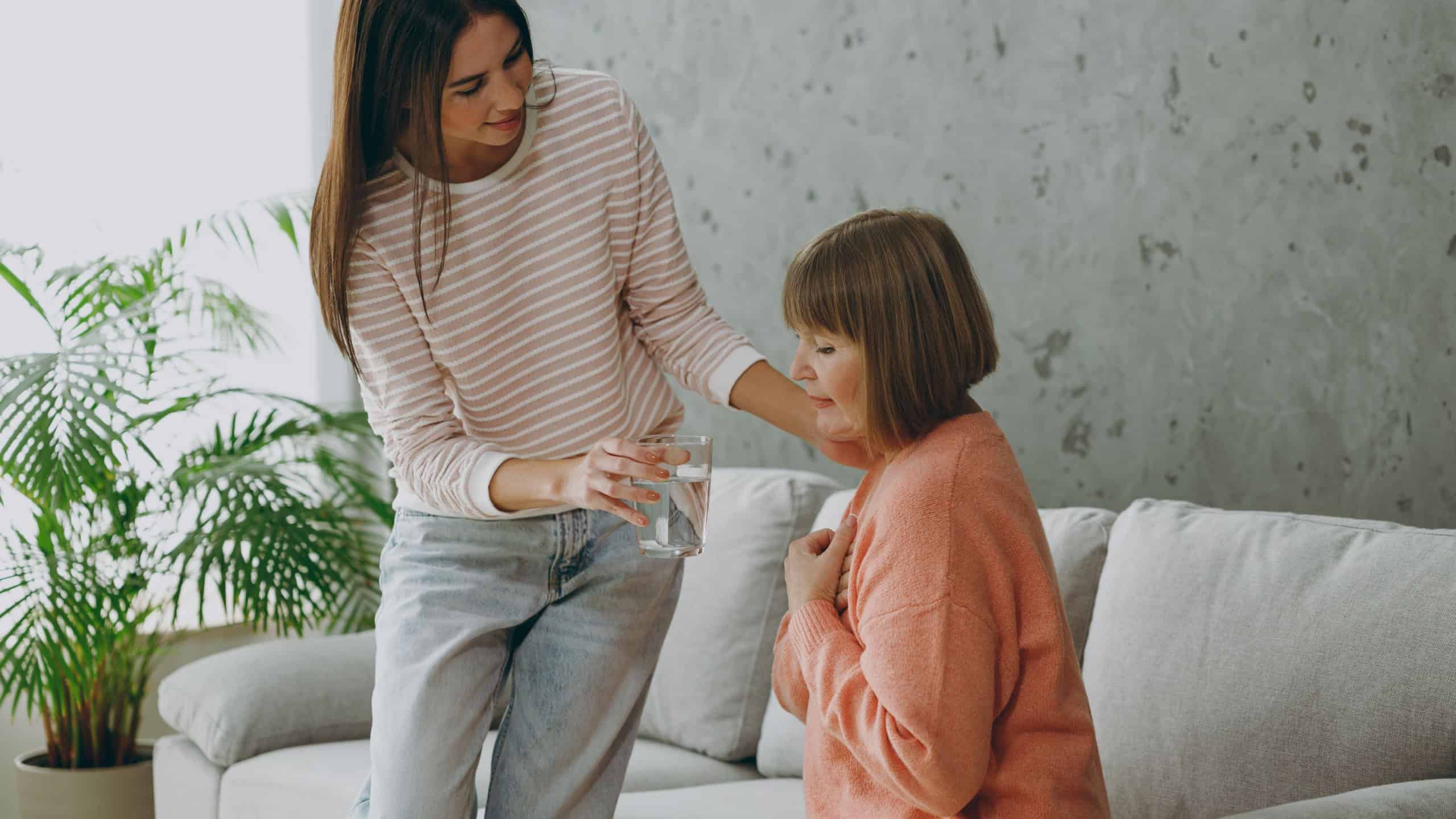 Two caring adult women mature mom young kid wear casual clothes give glass of water to drink sit on gray sofa couch stay at home flat rest relax spend free spare time in living room. Family concept