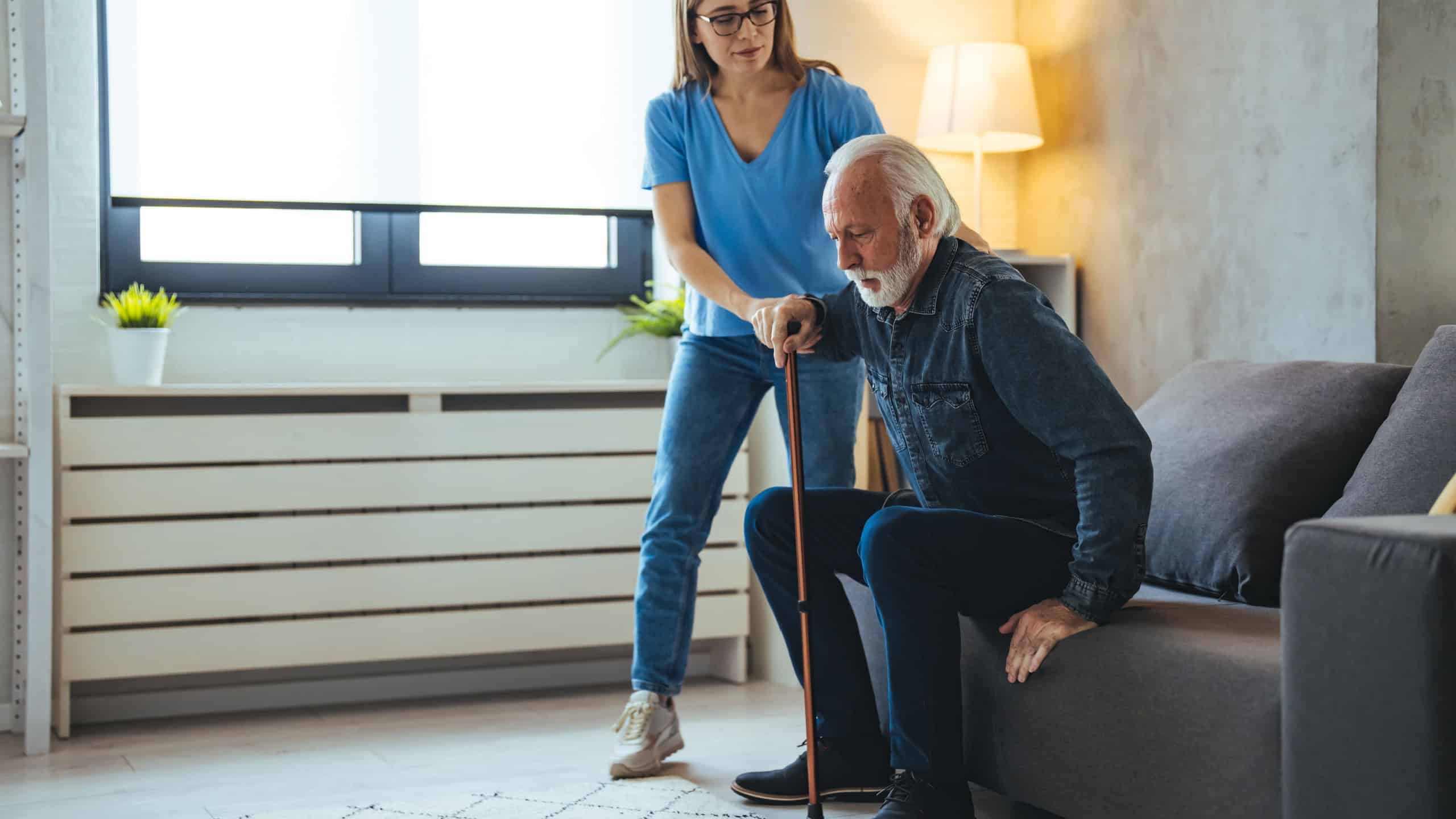 Smiling senior man with female healthcare worker. Home carer supporting old man to stand up from the chair at care home.. Female caregiver helping senior man get up from couch in living room - Indoors