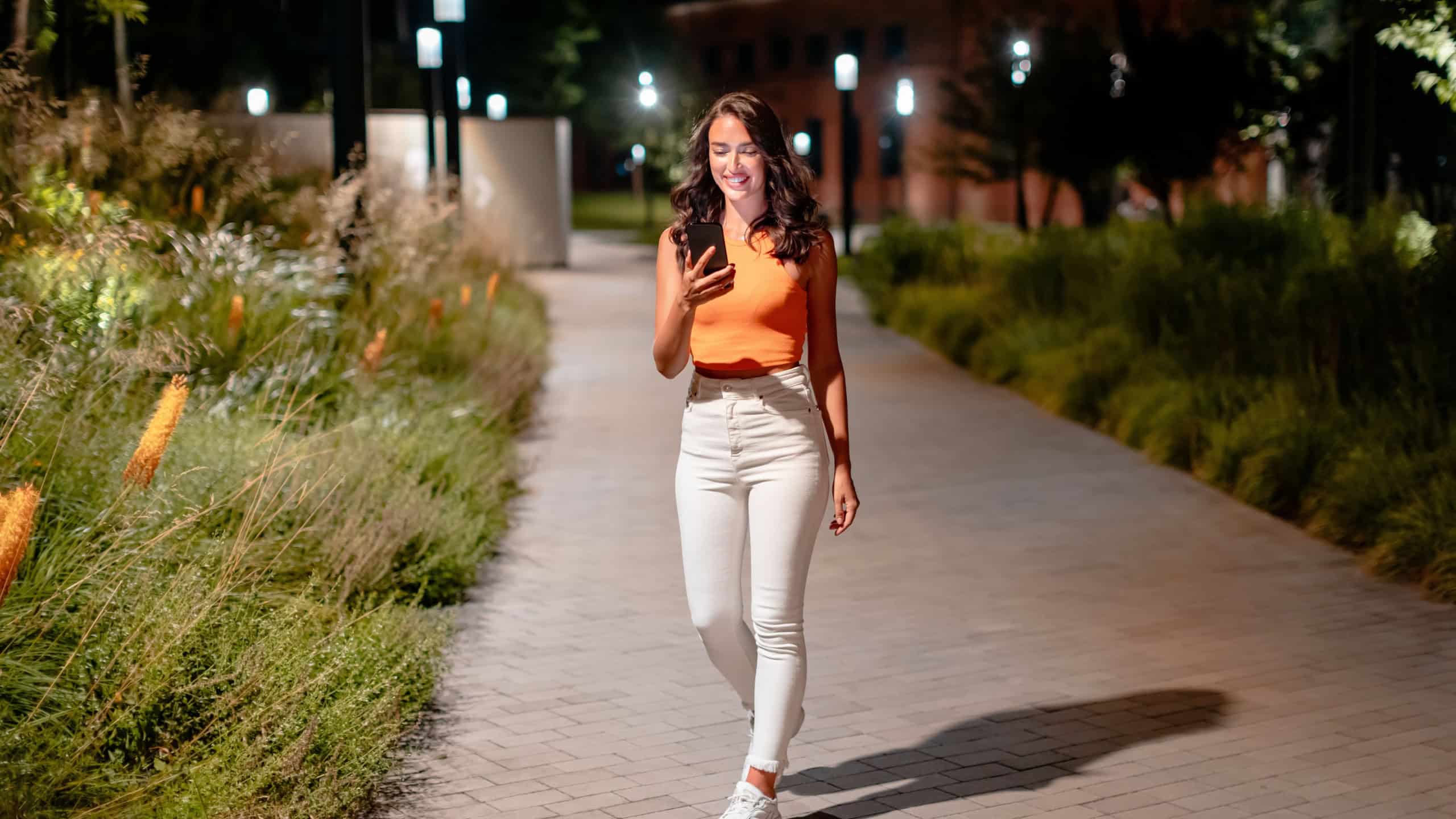 Full length shot of young european woman in casual outfit walking on the city street at night and using cellphone, texting or browsing online maps app, free space