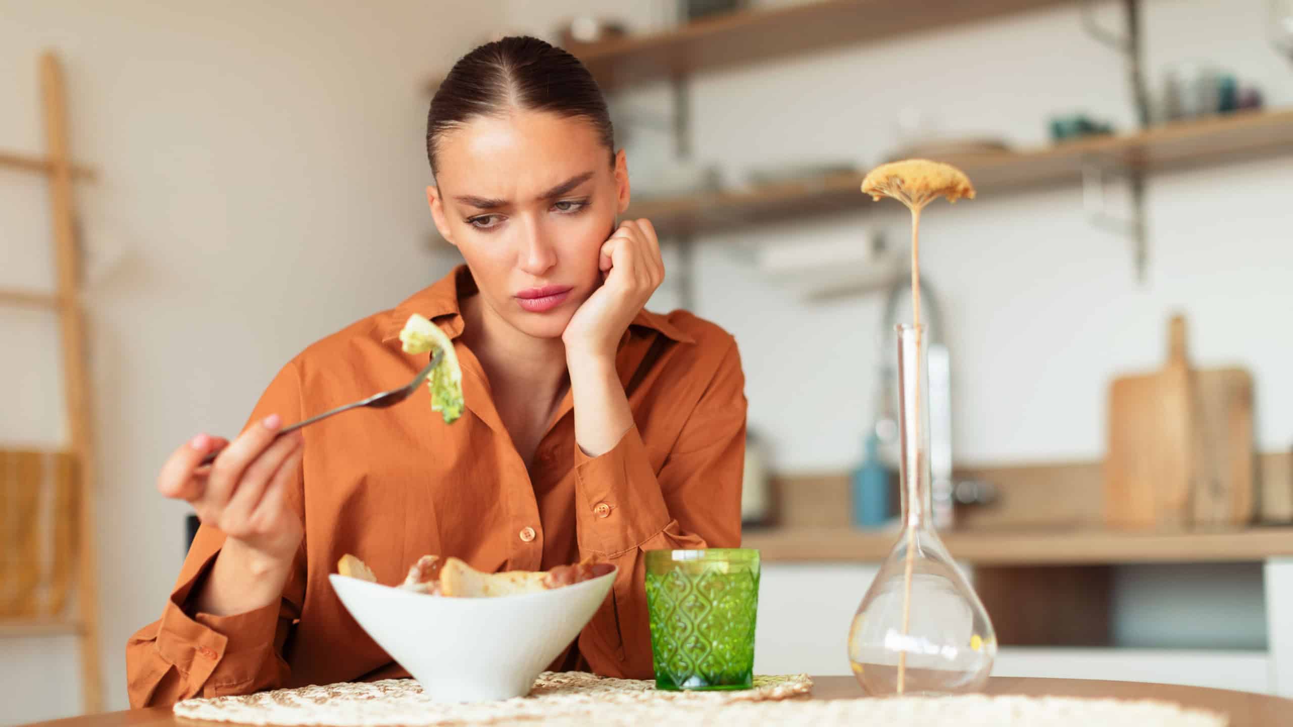 Sad young caucasian lady looking at her caesar salad and dont want to eat, sitting at kitchen interior at table, free space. Lack of appetite, mood swings