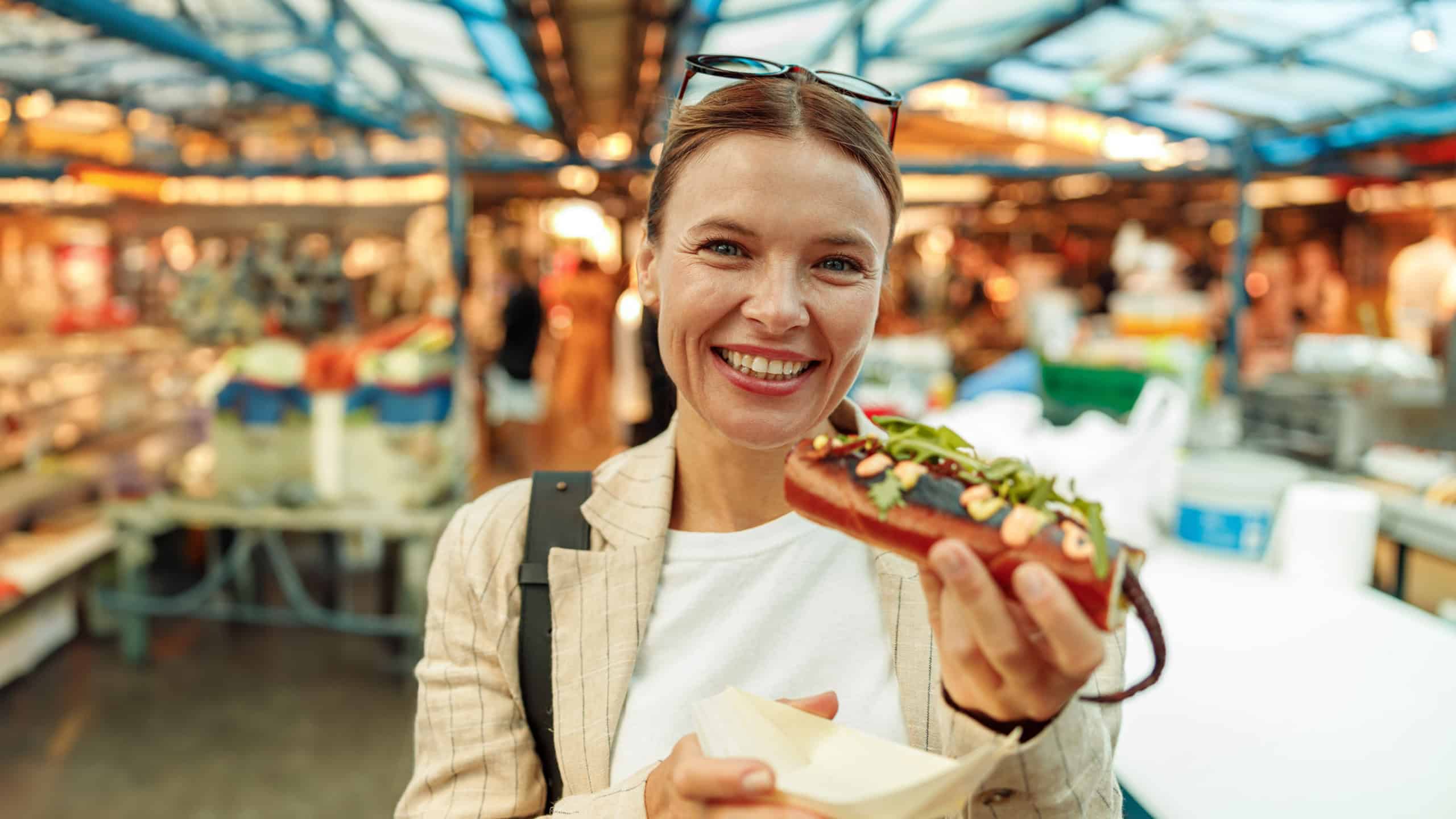 Smiling mature woman in eyeglasses standing with takeaway fast food hot dog on food market
