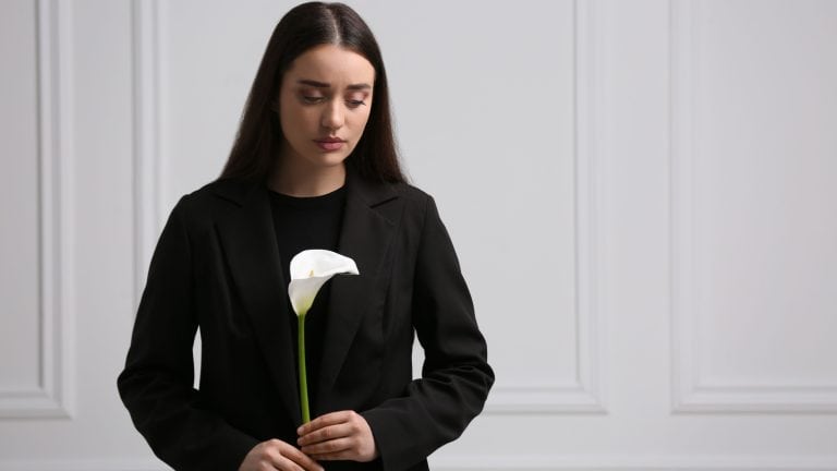 Sad woman with calla lily flower near white wall, space for text. Funeral ceremony