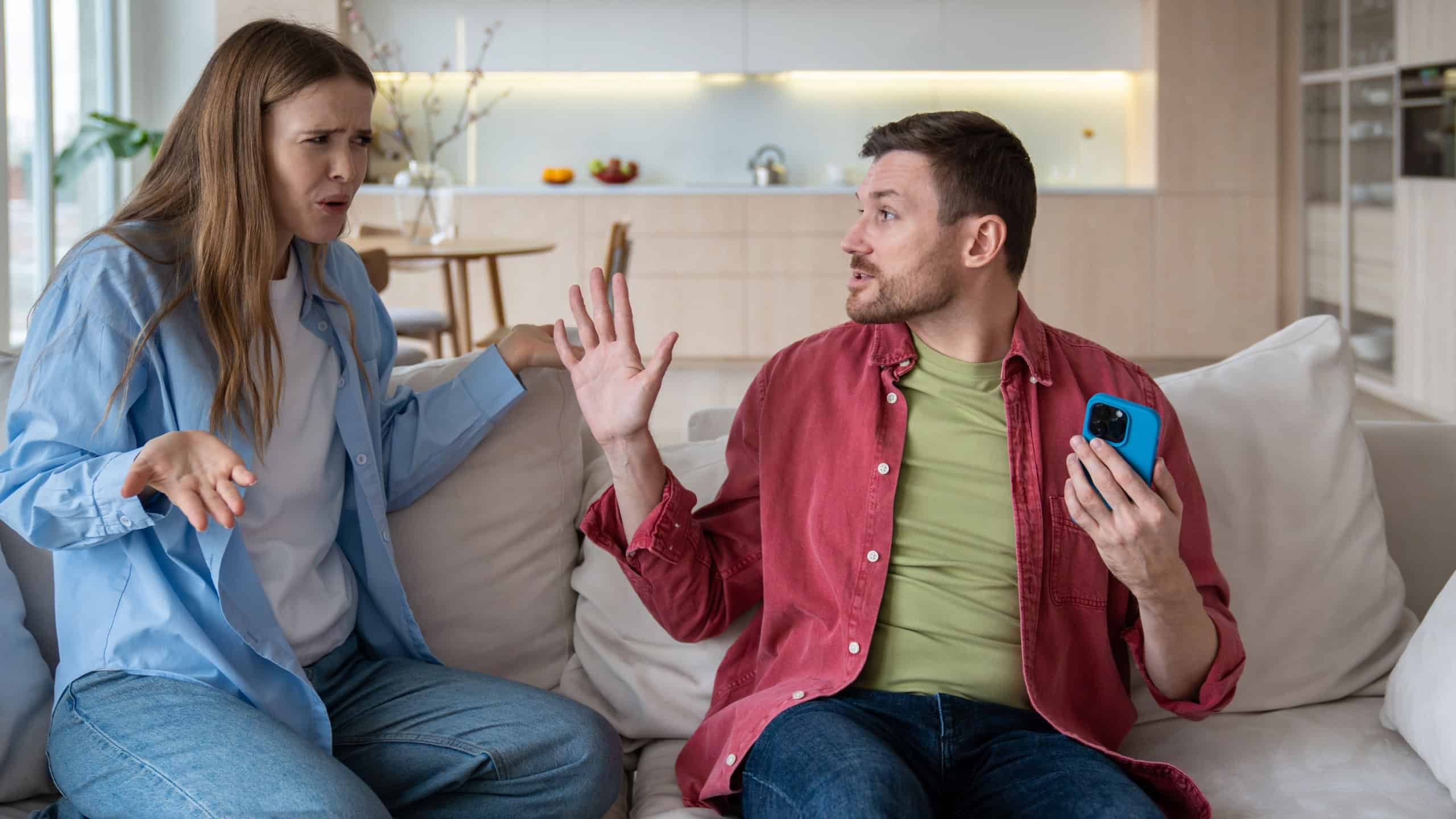 Jealous angry husband demands password from wife mobile phone sitting on couch at home. Family quarrel, scandal of two spouses. Tyrant man threatens woman. Betrayal infidelity mistrust relations.
