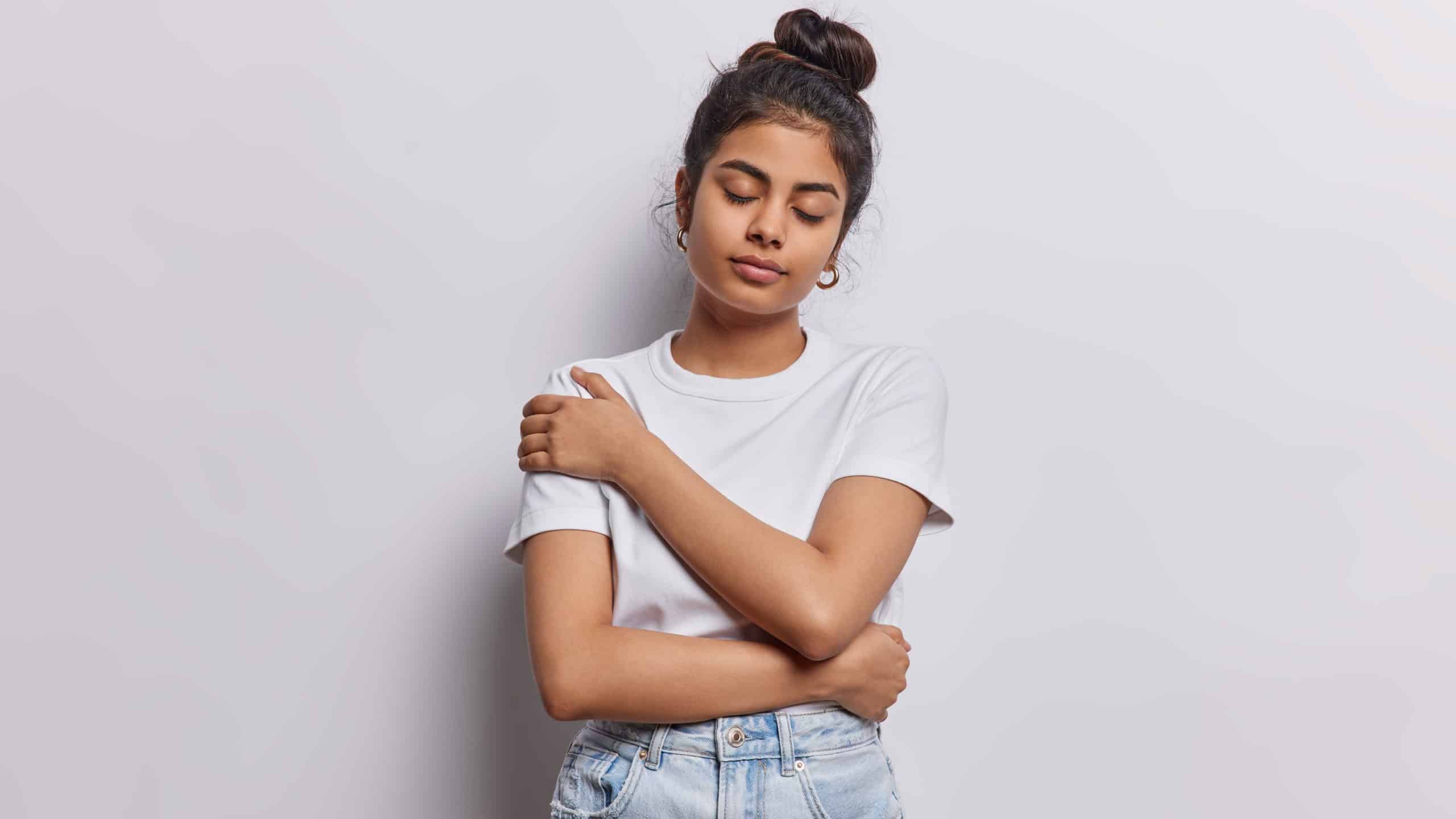 I love myself. Calm dark haired Iranian woman stands indoor alone embraces own body recalls pleasant memories wears casual t shirt and jeans isolated over white background has romantic mood.