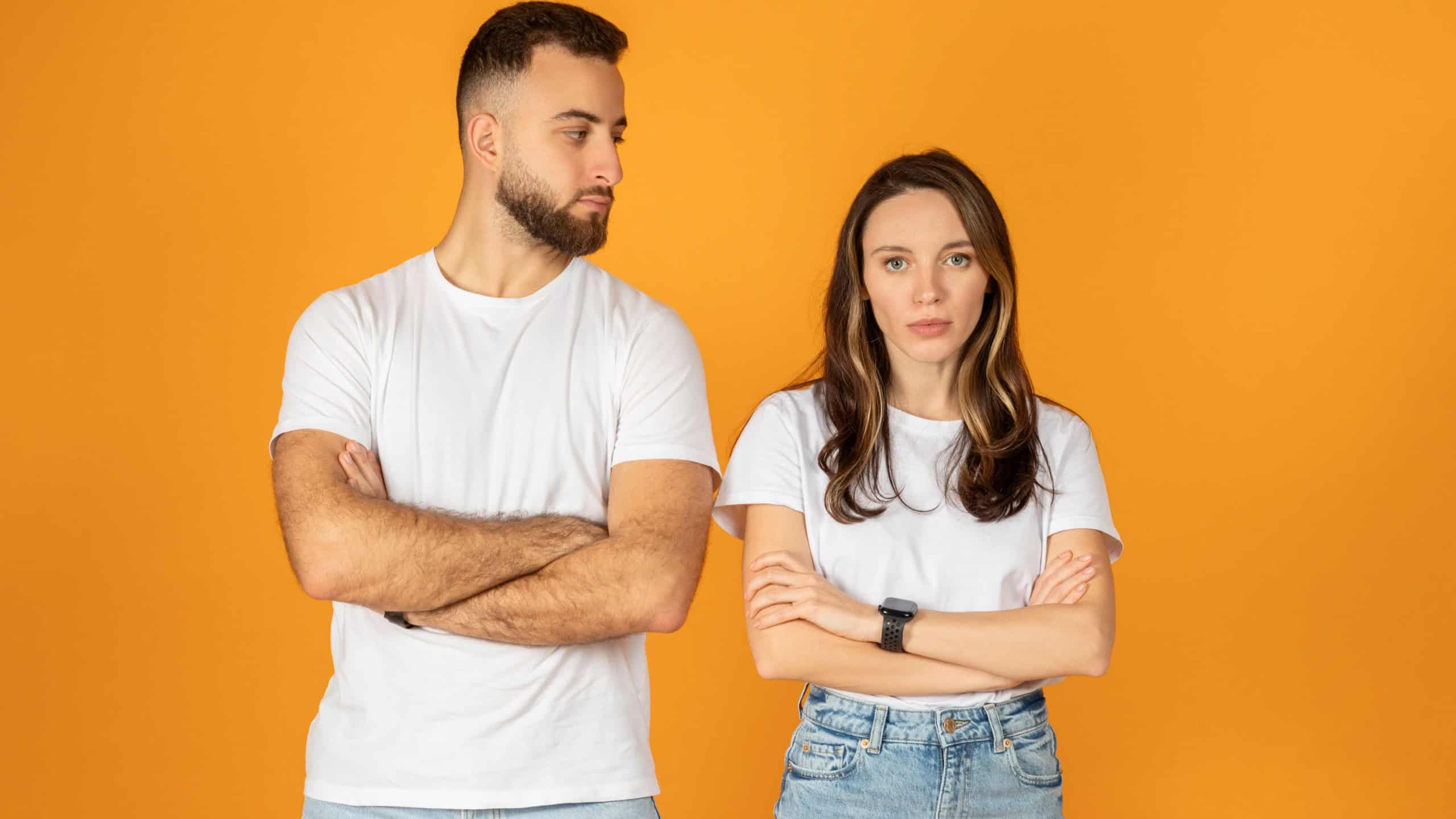 A skeptical sad european young man and woman stand with arms crossed, looking at each other with questioning expressions, against orange background, studio, relationship problems