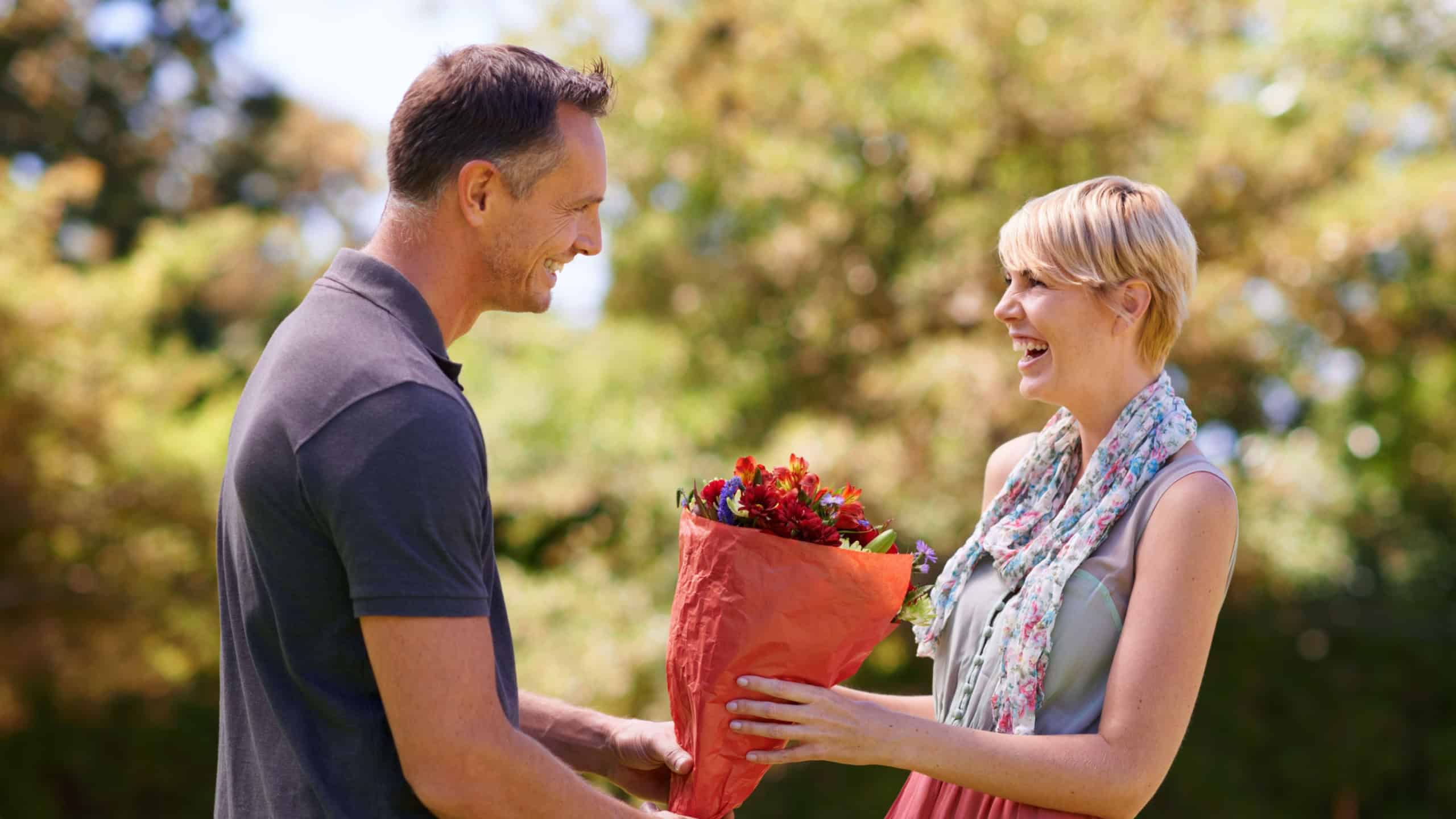Smile, couple and present of flowers in nature outdoor, bonding and date on valentines day. Bouquet, man and woman with gift for love, care and romantic connection of people laughing at park together
