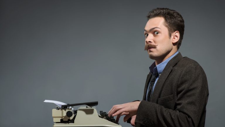 Just inspired . Young nerd author typing something at the typewriter and looking at camera while sitting at his working place against grey background