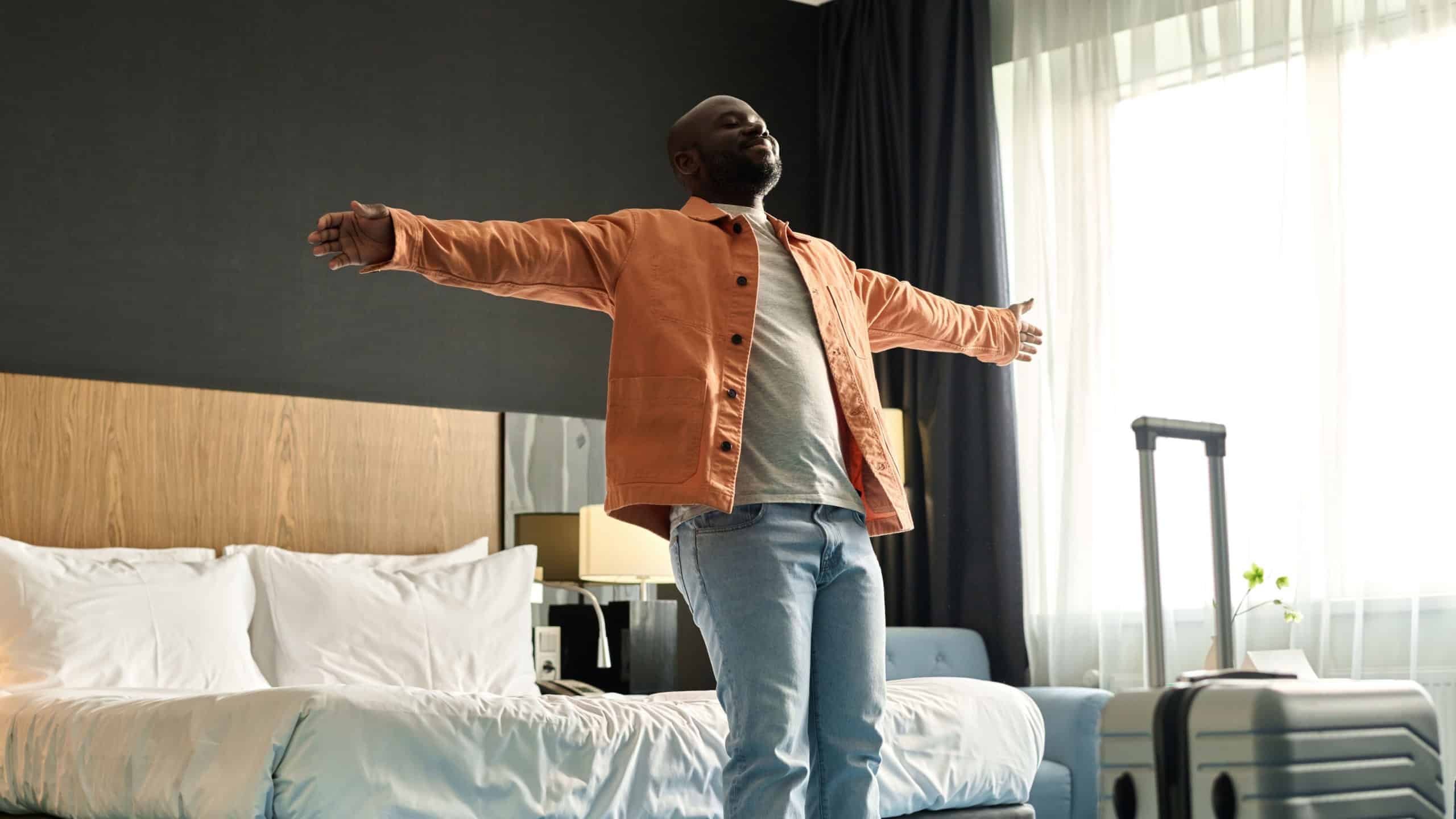Portrait of joyful Black man falling onto bed with arms spread arriving in luxury hotel copy space