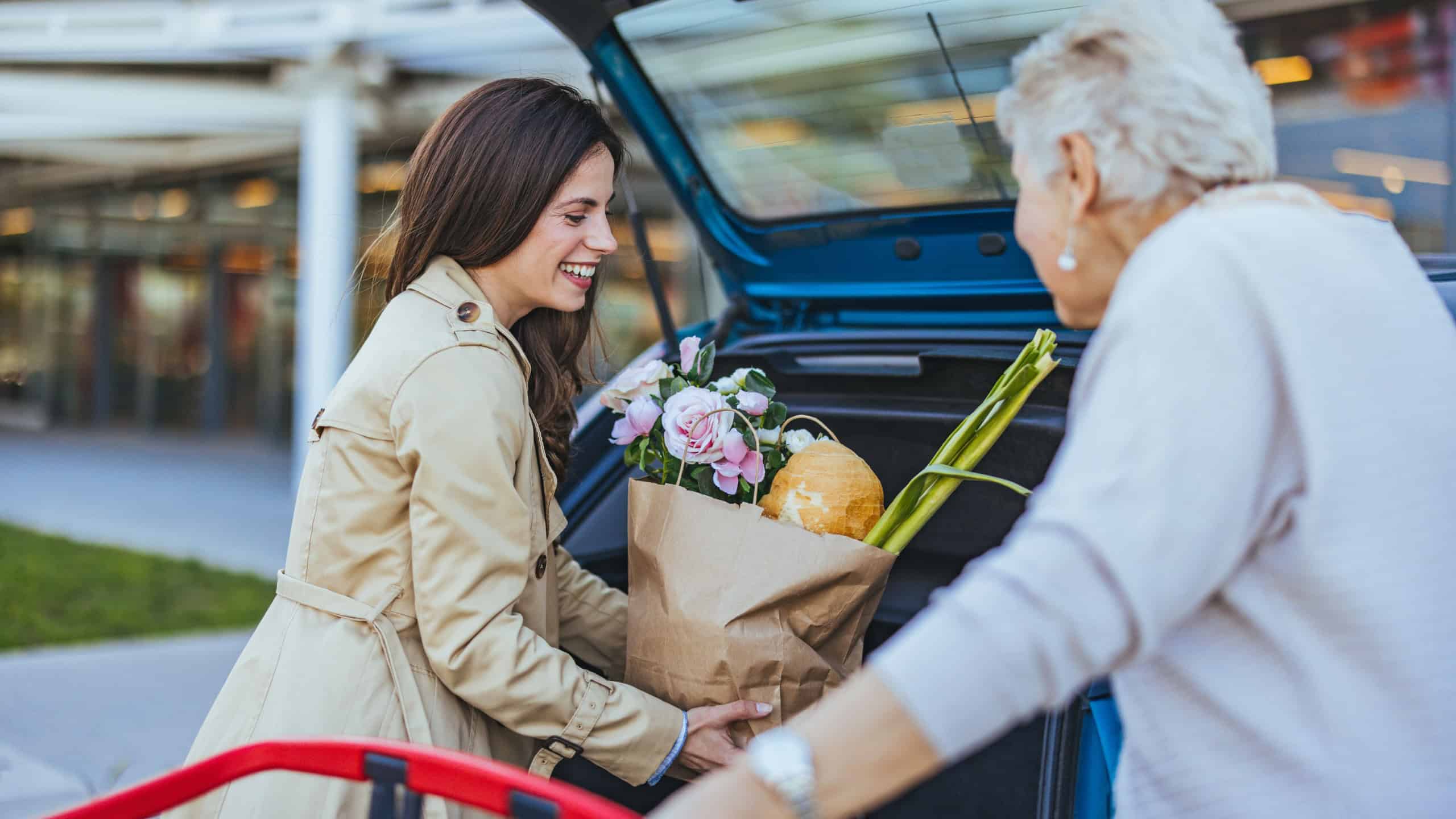 A joyful Caucasian woman assists her elderly mother in transferring grocery bags from a trolley to the open trunk of a vehicle in a parking lot.