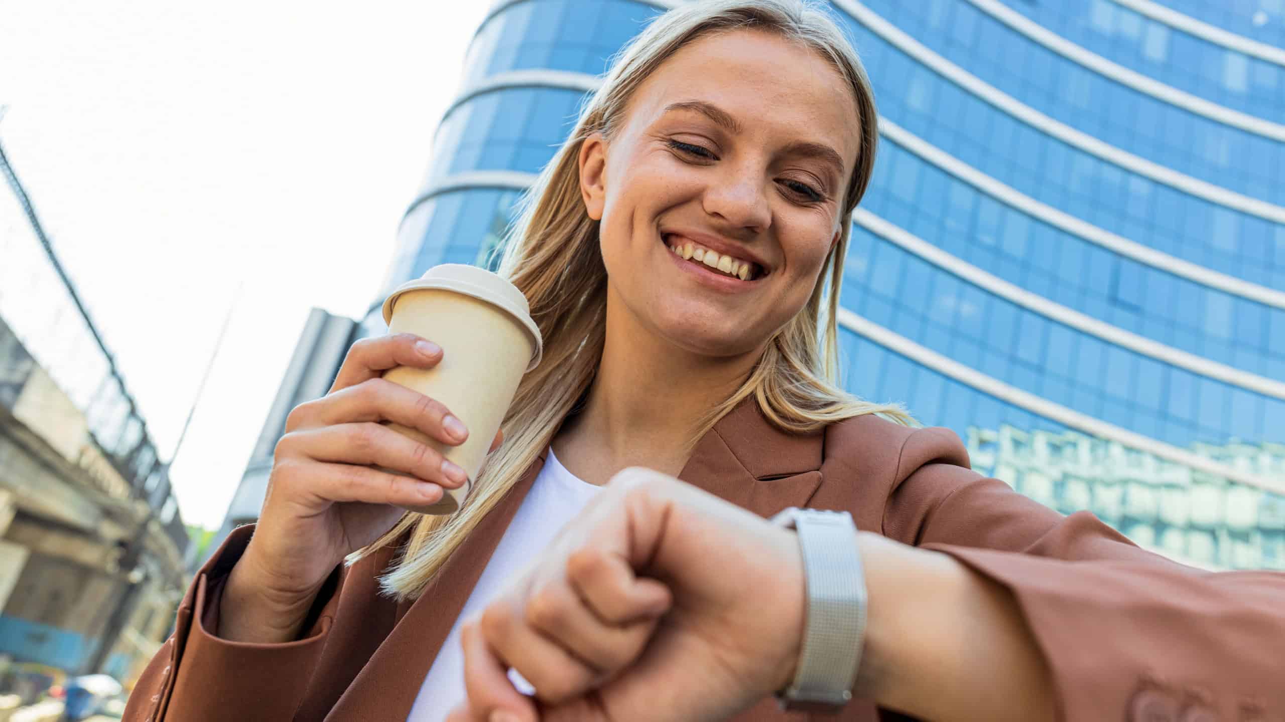 Smiling business woman with paper cup looking at her watch against the backdrop of business center