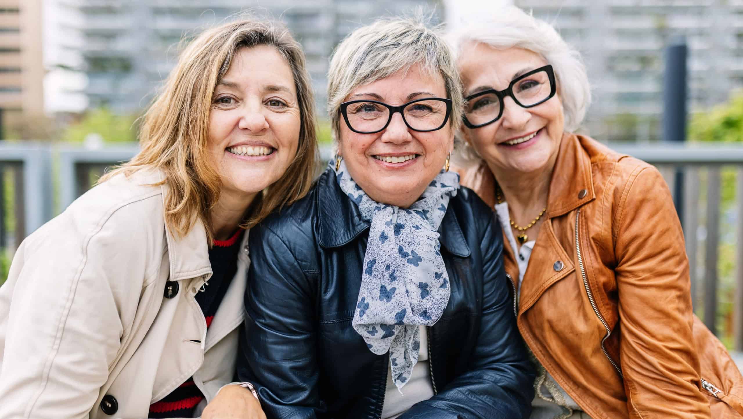 Portrait of three mature retired women smiling at camera sitting outside. Small group of mature females having fun at city street. Elderly friendship concept.