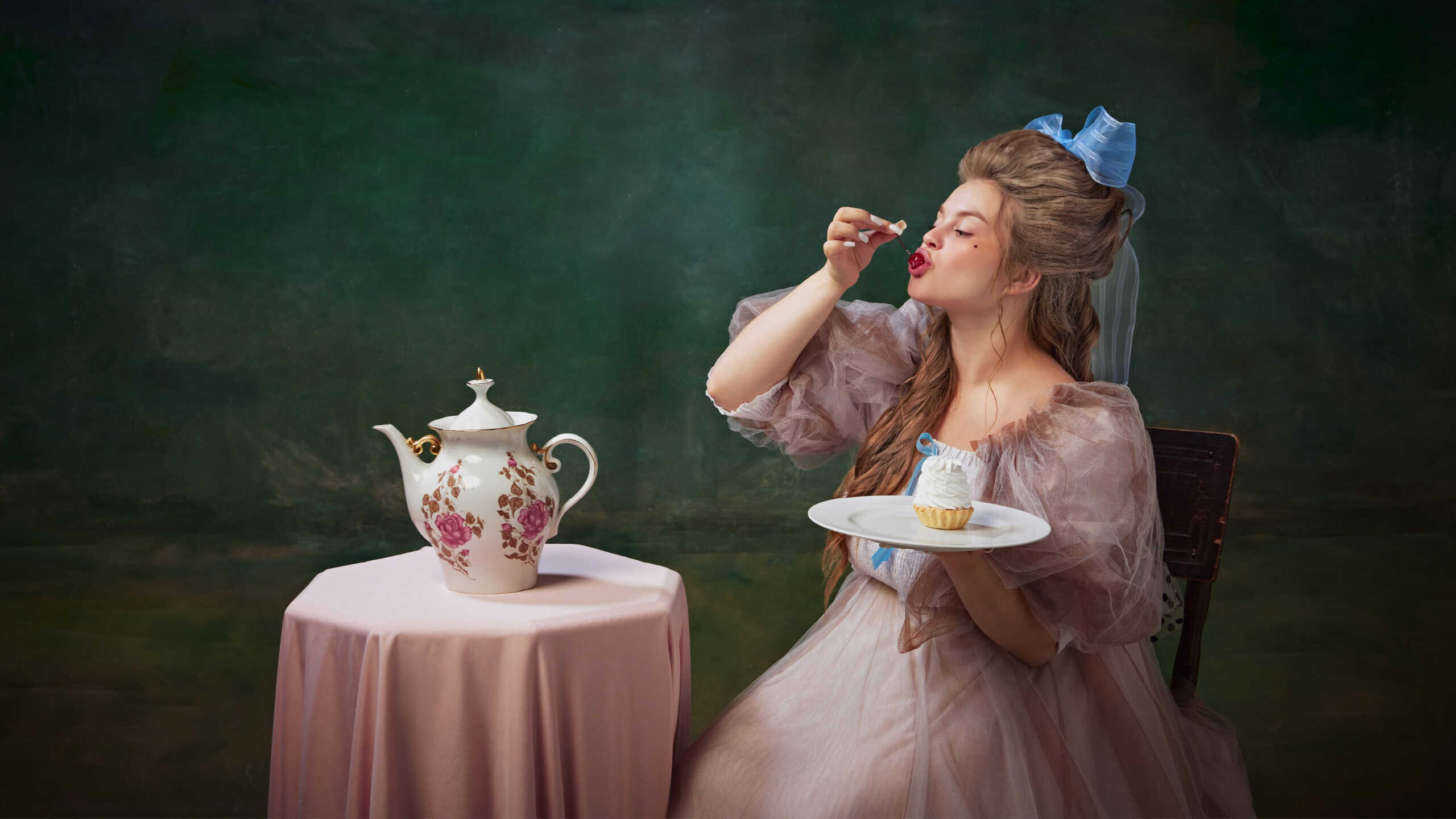 Portrait of beautiful young woman in elegant period attire sitting at table and eating cherry from cake against vintage green background. Concept of history, baroque art, comparison of eras