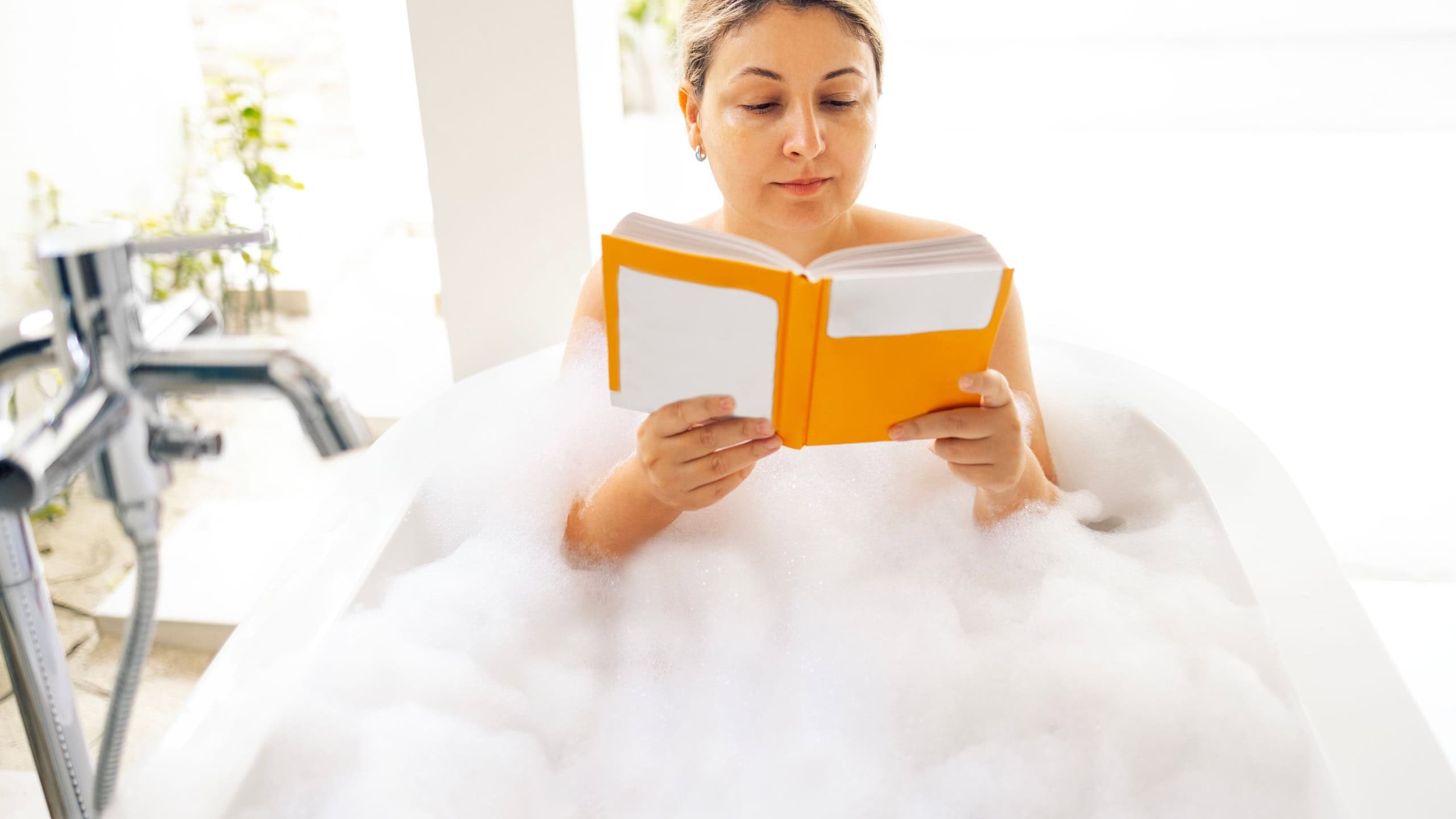 A young woman holds a book and takes a bubble bath