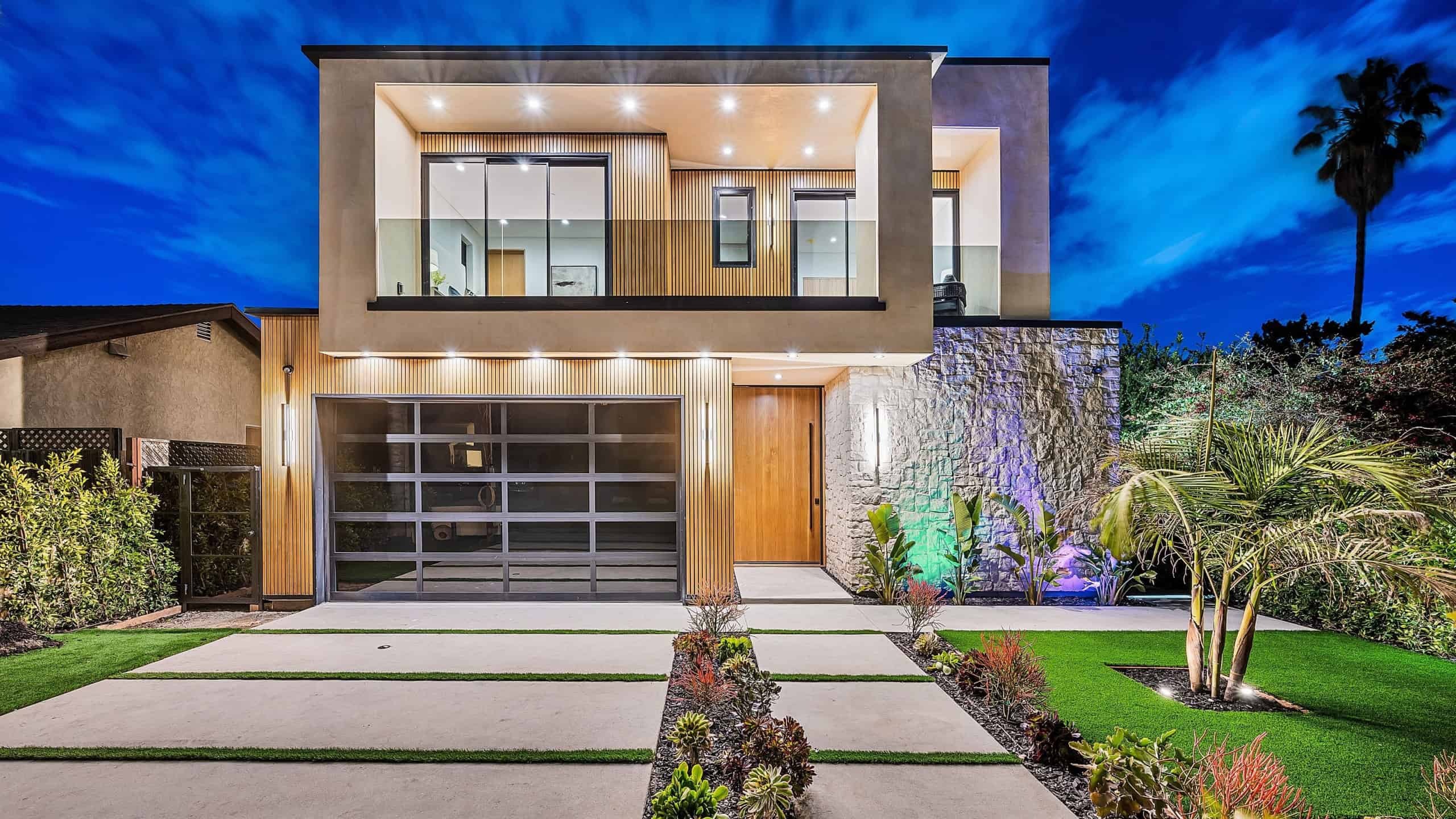 Modern two-story house with large windows and a landscaped front yard, illuminated at night with a clear blue sky.