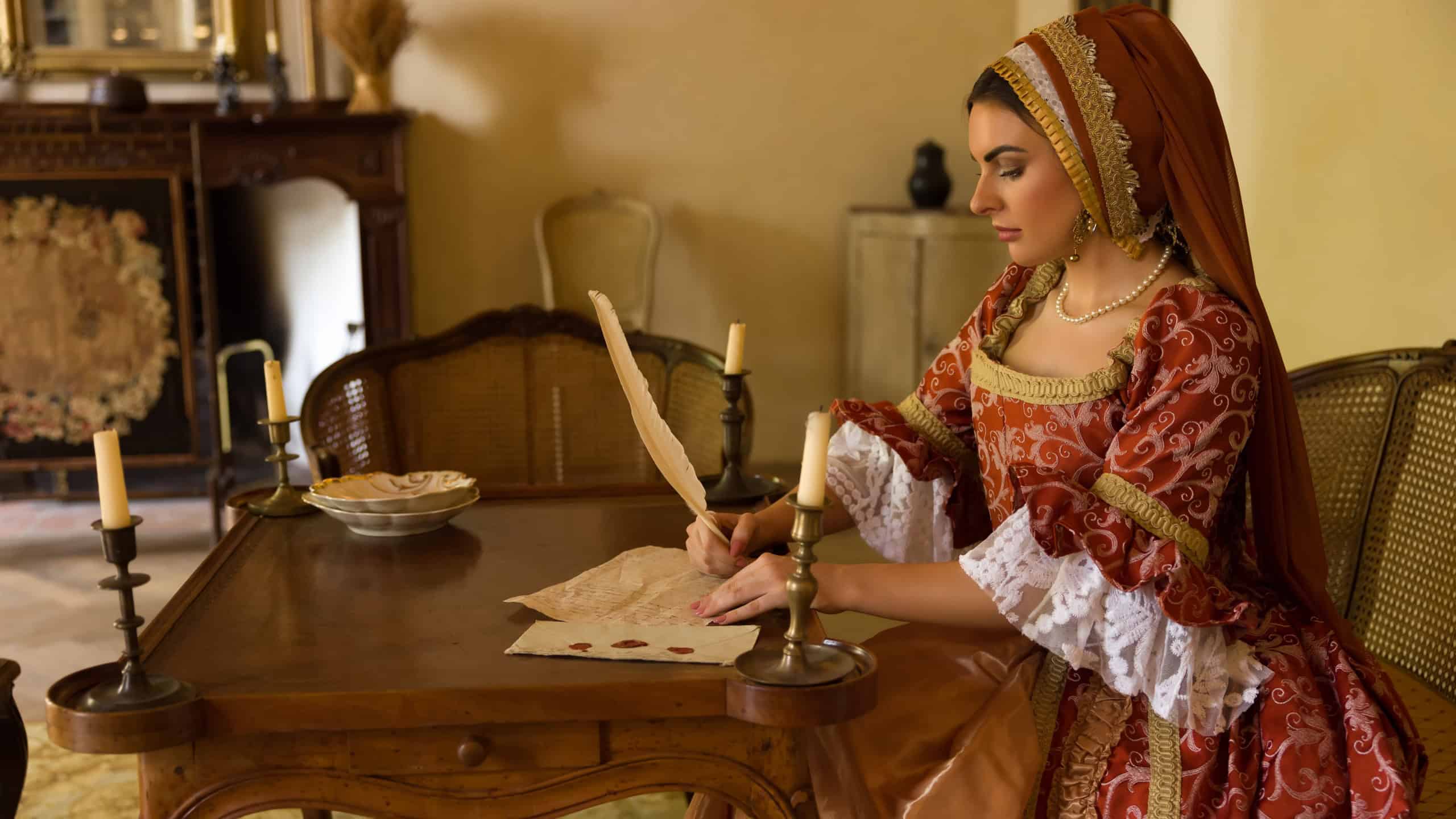 Woman wearing renaissance gown and French hood writing a letter at her table.