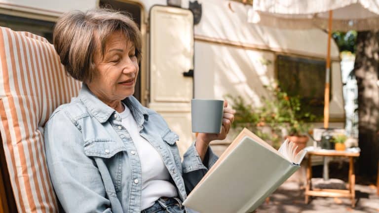 Relaxed caucasian senior old elderly woman grandmother granny reading book, learning near trailer porch, traveling by motor home, caravanning by camper van