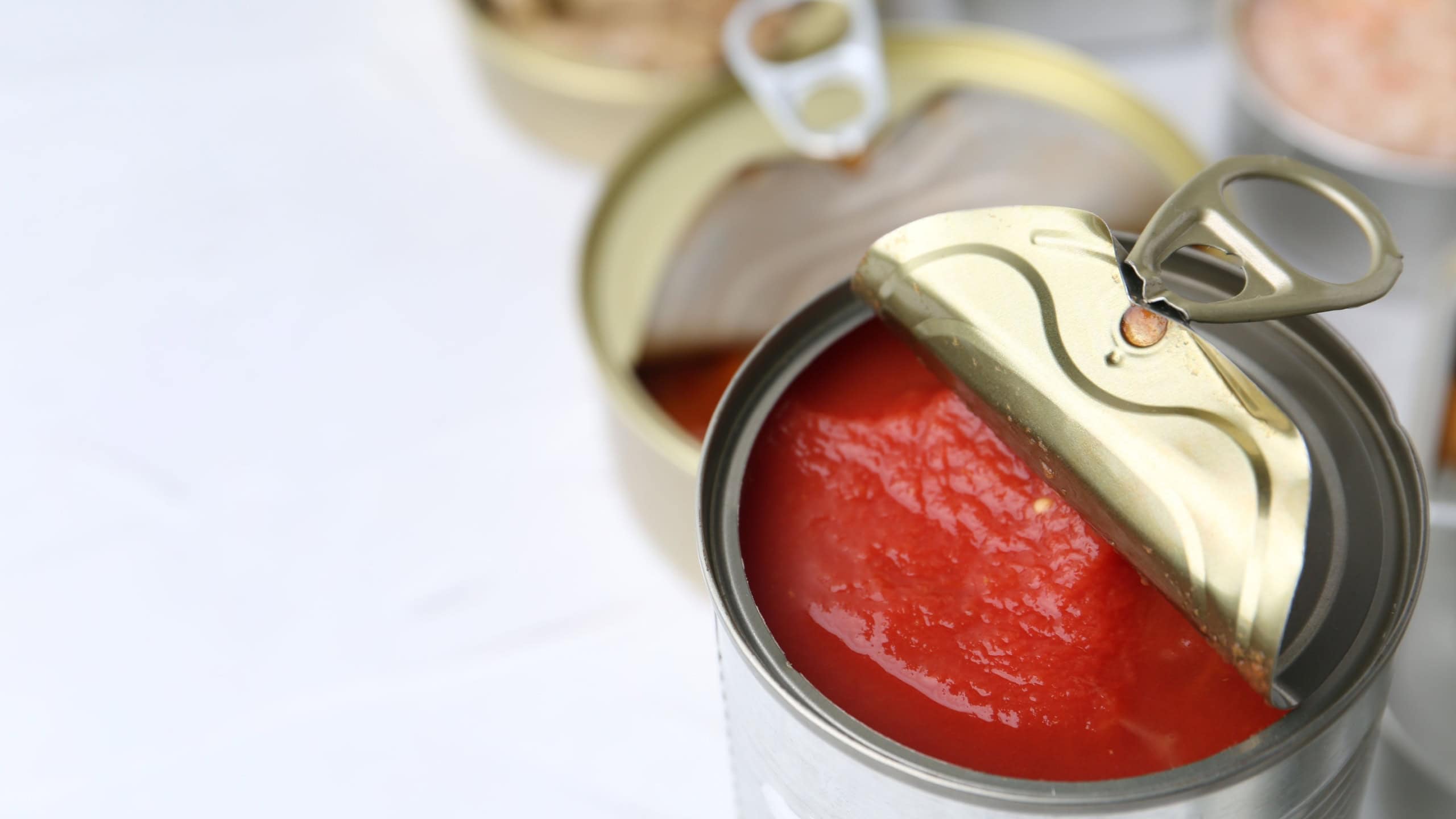 Open tin cans with different preserved products on white table, closeup. Space for text
