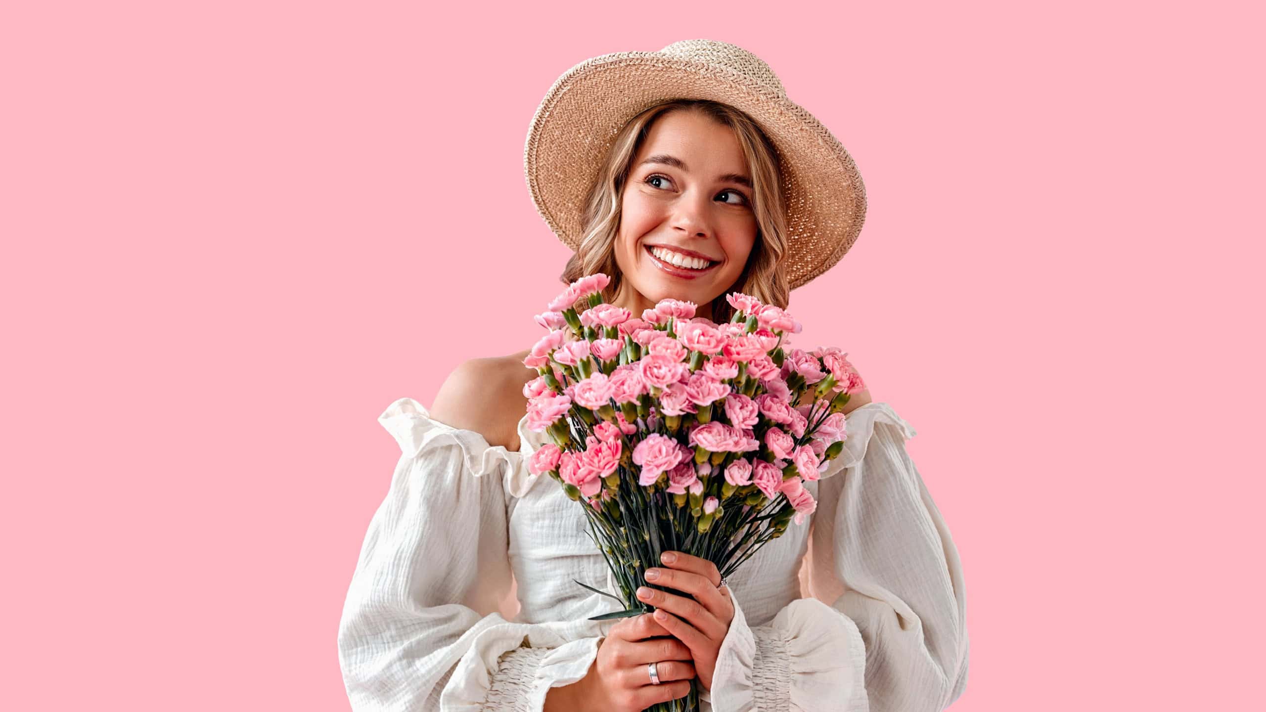 Smiling happy dreamy girl in straw hat and trendy dress holding bouquet of pink carnations and looking aside while standing on pink background. Stylish young girl with delicate pink flowers.