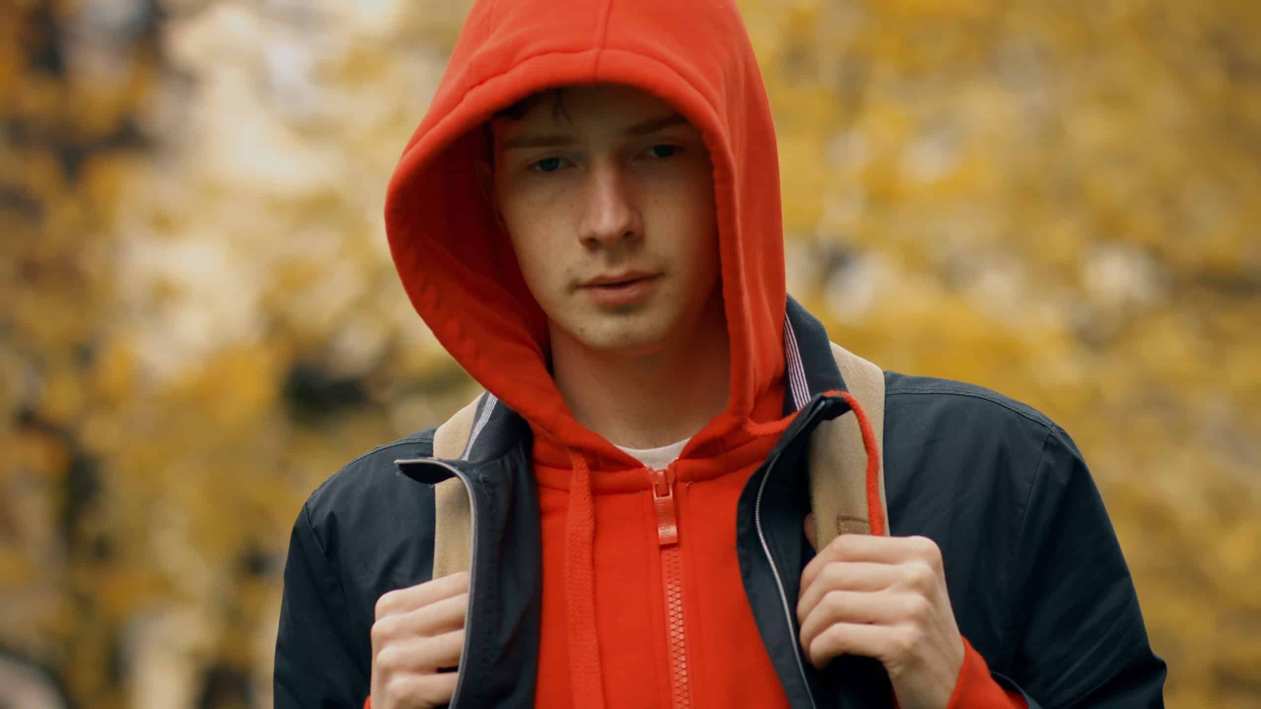 Sad caucasian teenager boy with backpck outdoors in autumn park. Close up of teen kid in red hoodie and black jacket looking at camera