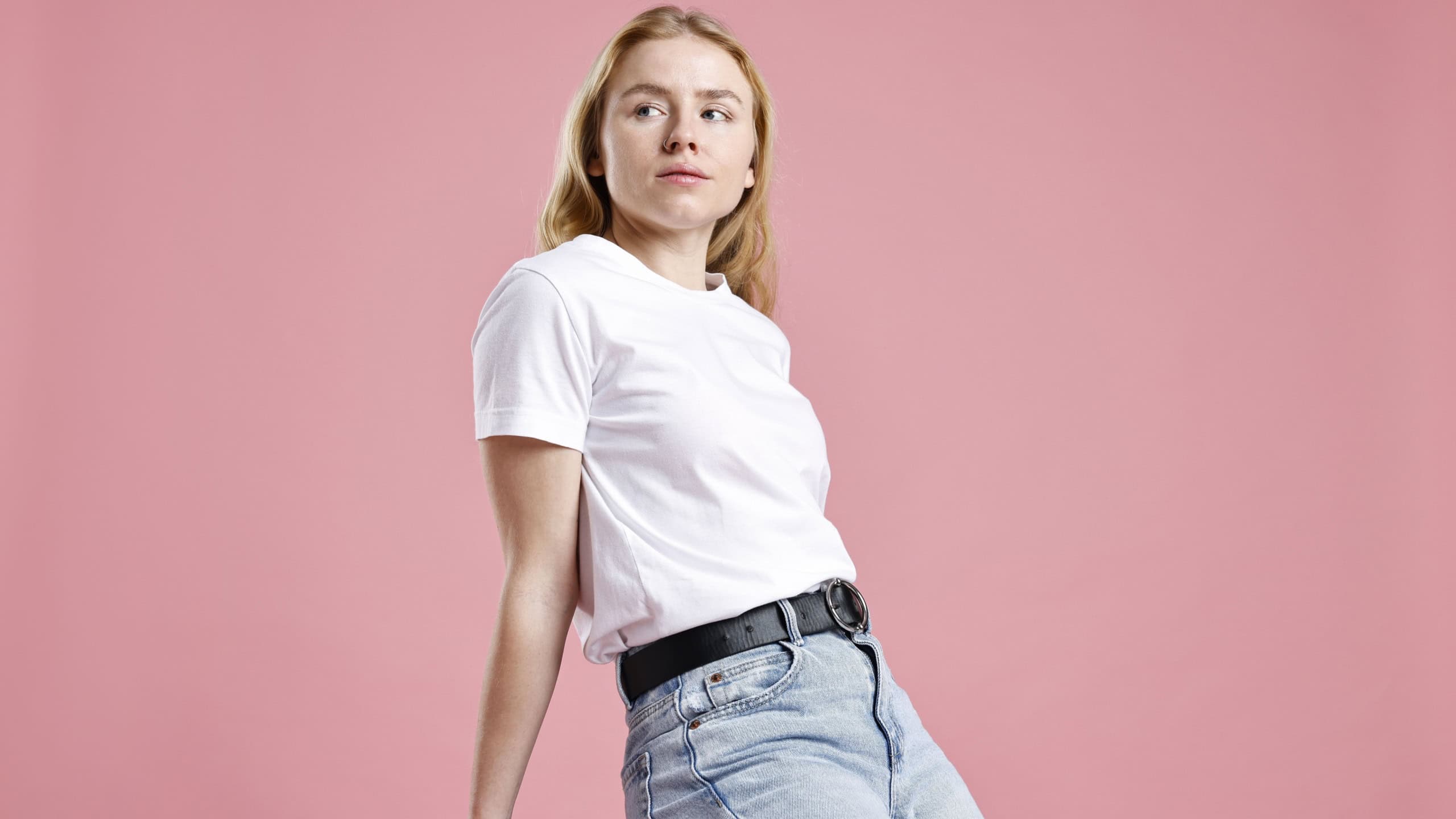 Woman in stylish jeans sitting on stool against pink background, low angle view